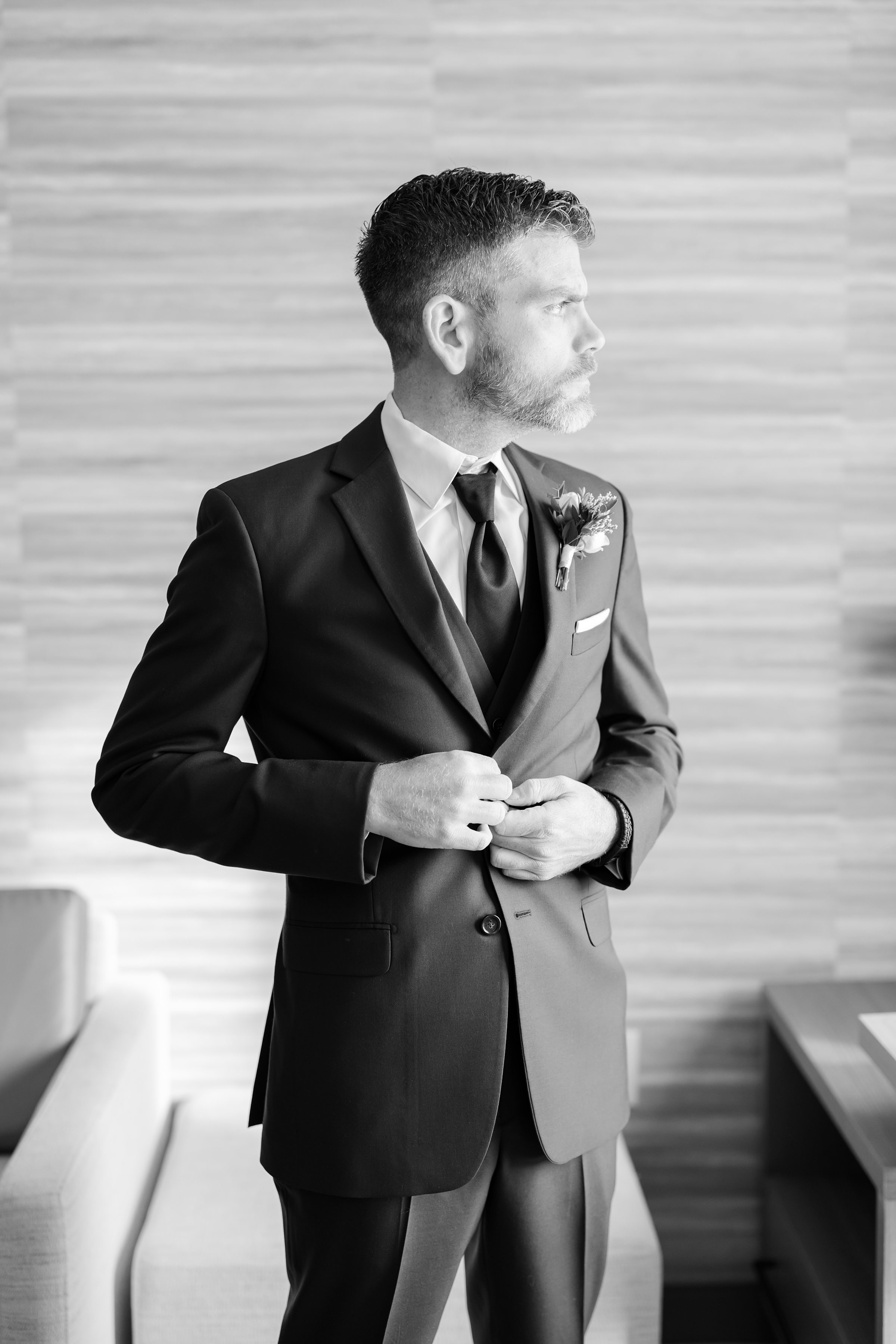 Groom adjusting his suit jacket while looking off to the side indoors.