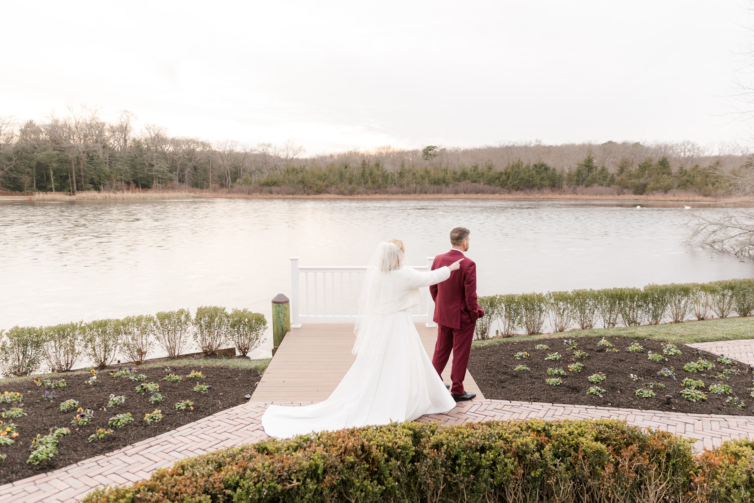 Bride gently reaches for her groom as they stand together by a peaceful lakeside, her flowing white gown trailing behind while he looks out over the water in a quiet, intimate moment.