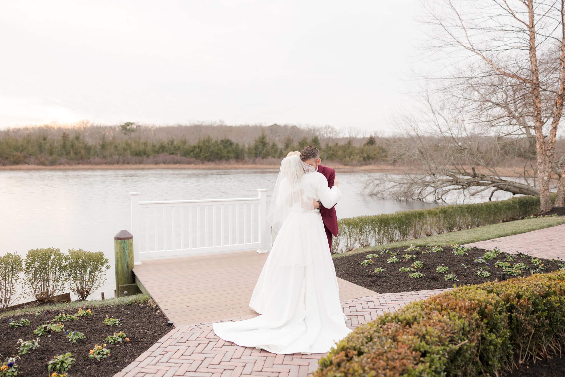 Bride and groom embrace by a quiet lakeside, her gown flowing behind them.