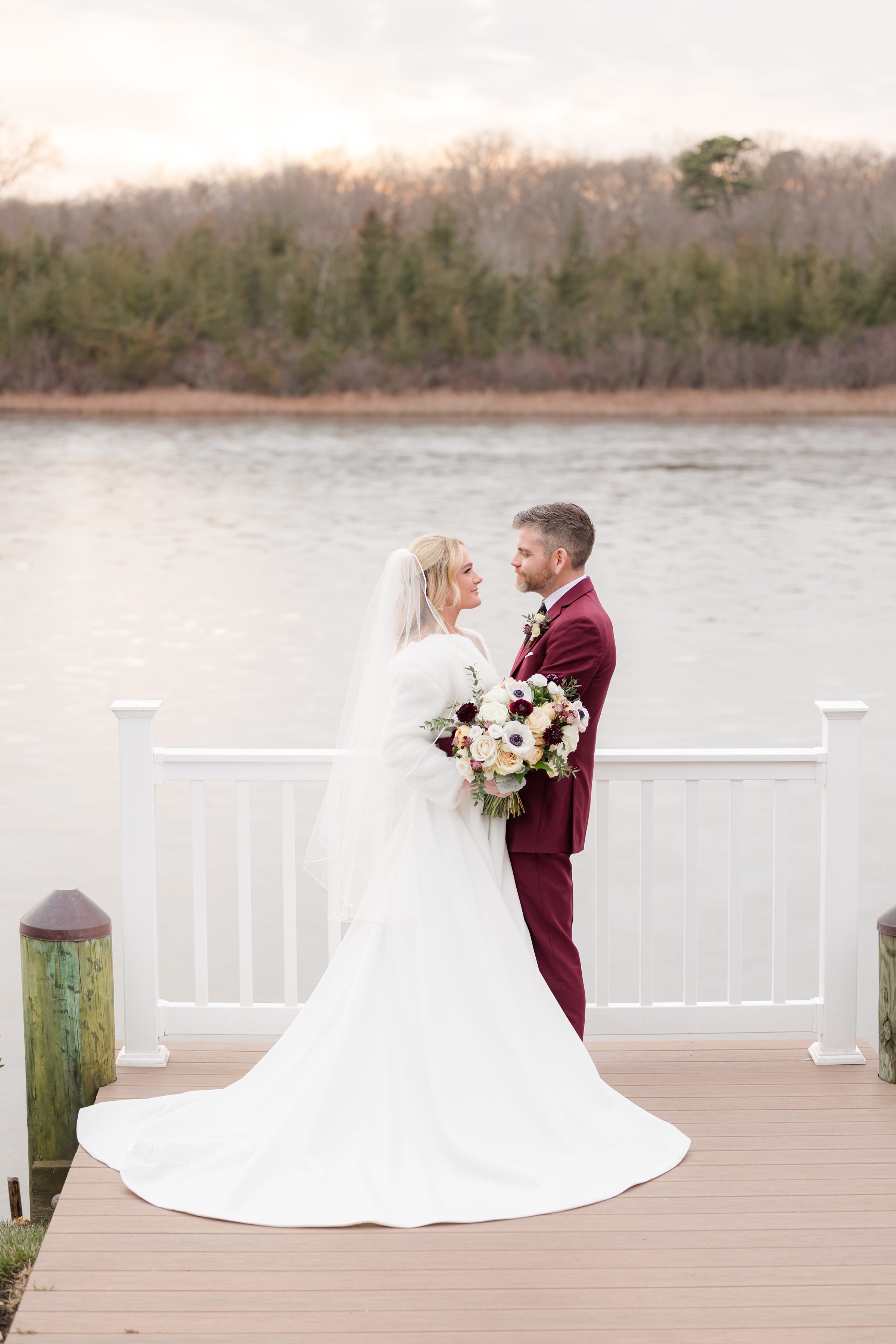 Bride and groom face each other by the water, holding a bouquet in a quiet, romantic moment.