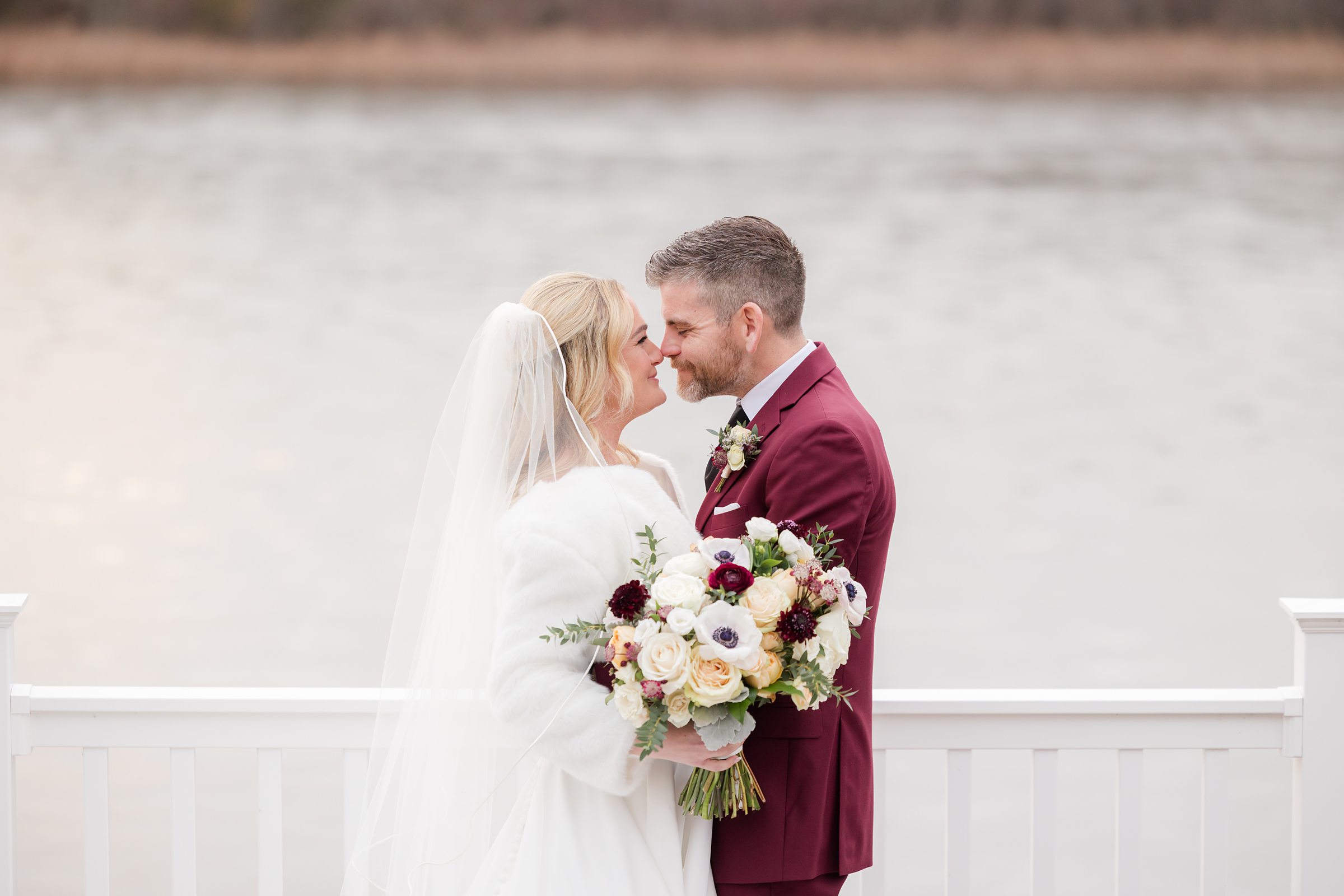 Bride and groom lean in close, smiling softly while holding a bouquet by the water.