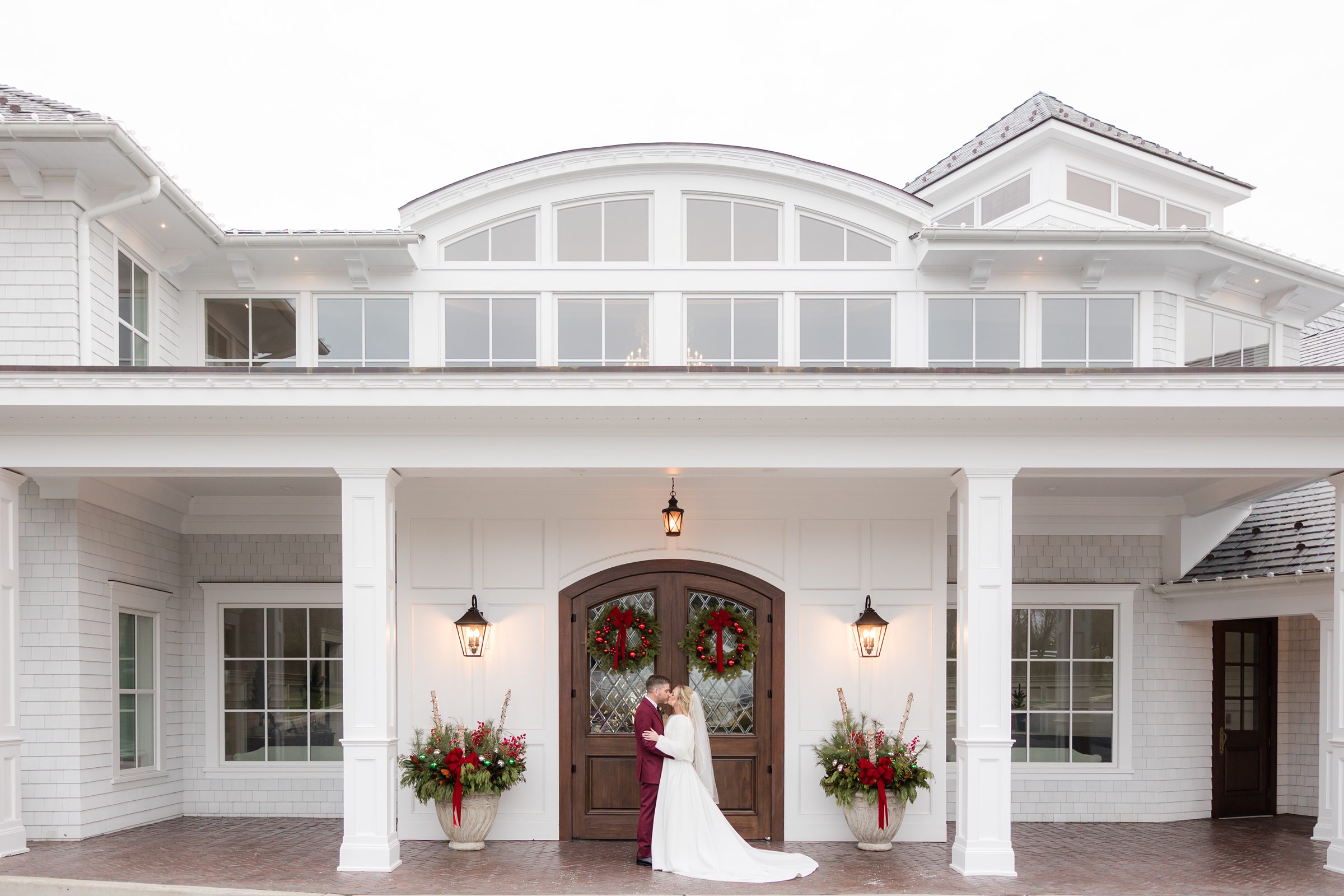 Bride and groom share a kiss in front of an elegant white venue with festive holiday décor.