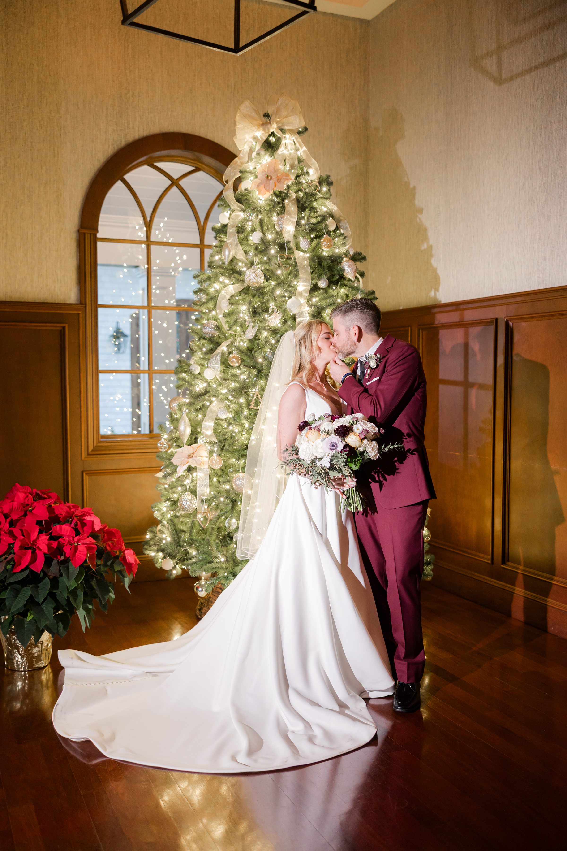 Bride and groom share a soft kiss in front of a glowing Christmas tree, surrounded by warm holiday light.