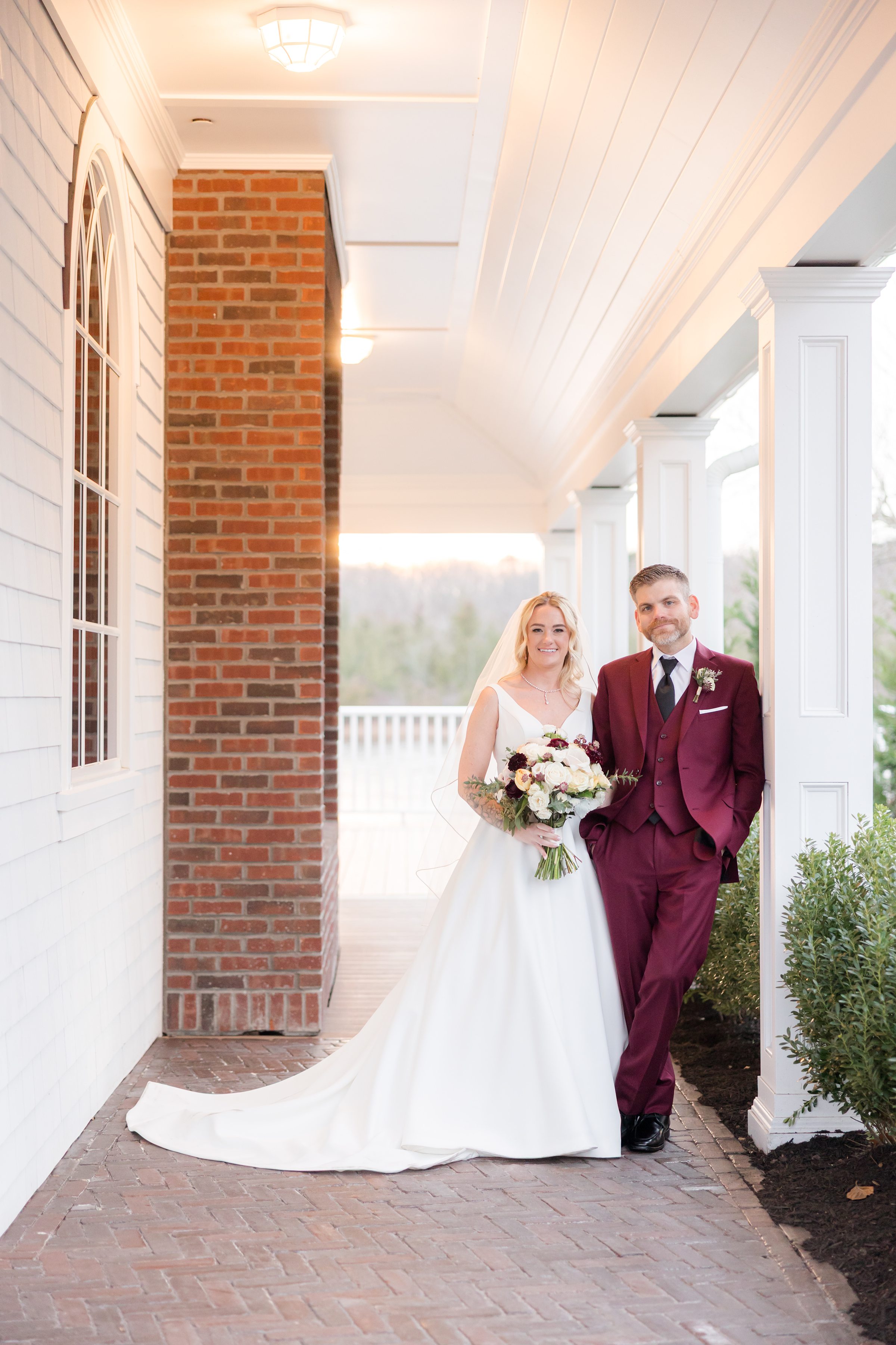 Bride and groom standing together on a covered porch, smiling softly as golden light surrounds them, her white gown flowing beside his deep burgundy suit.