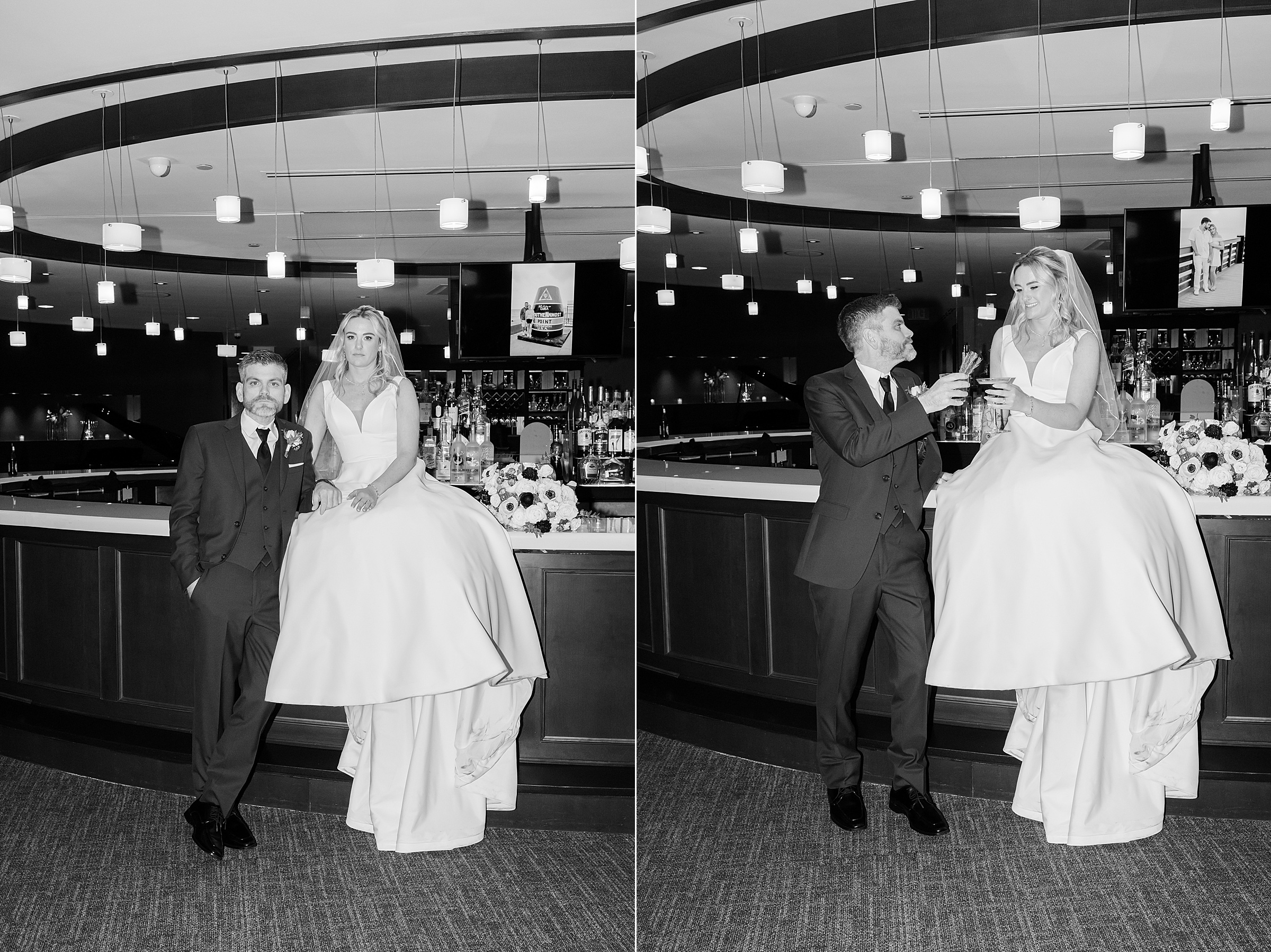 Bride in a full white gown sits on a bar counter while the groom stands beside her, both smiling and sharing drinks under soft, modern lighting in an intimate black-and-white reception moment.