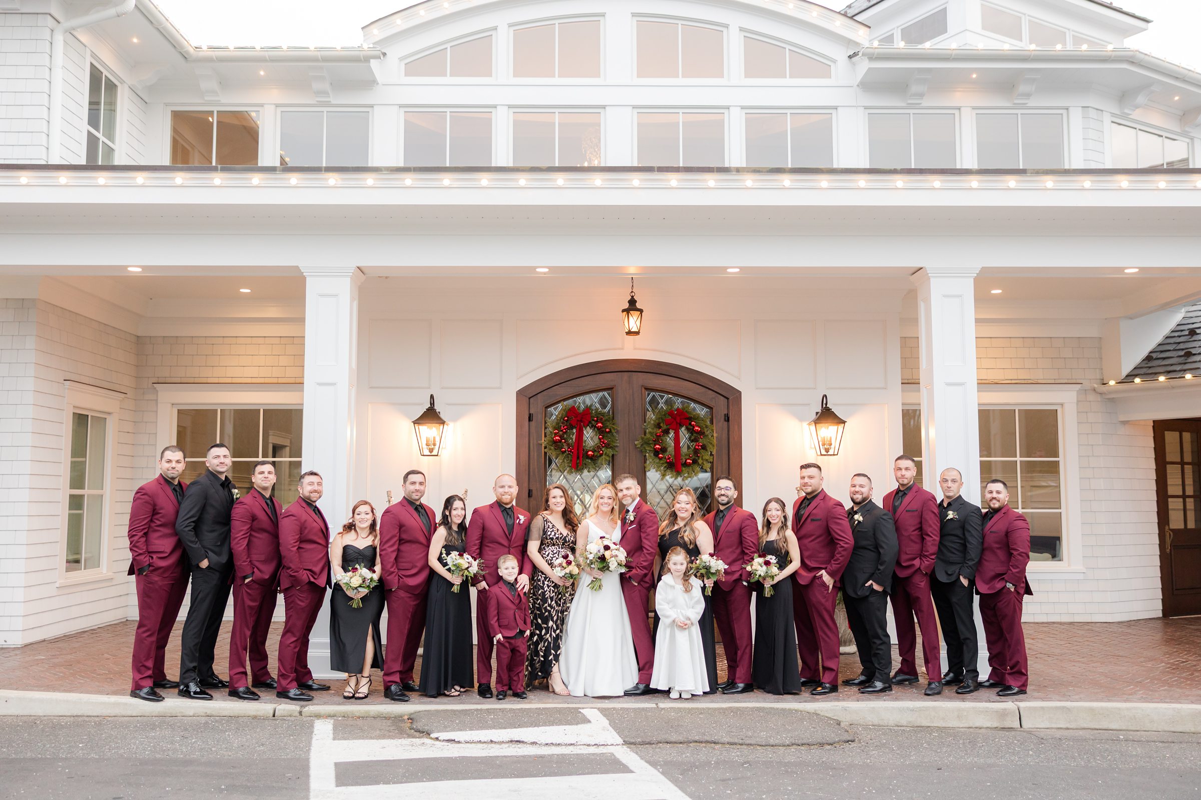Wedding party gathers in front of a white, elegant venue decorated with holiday wreaths, with the bride and groom centered and surrounded by family and friends in coordinated formal attire, capturing a joyful and celebratory group portrait.