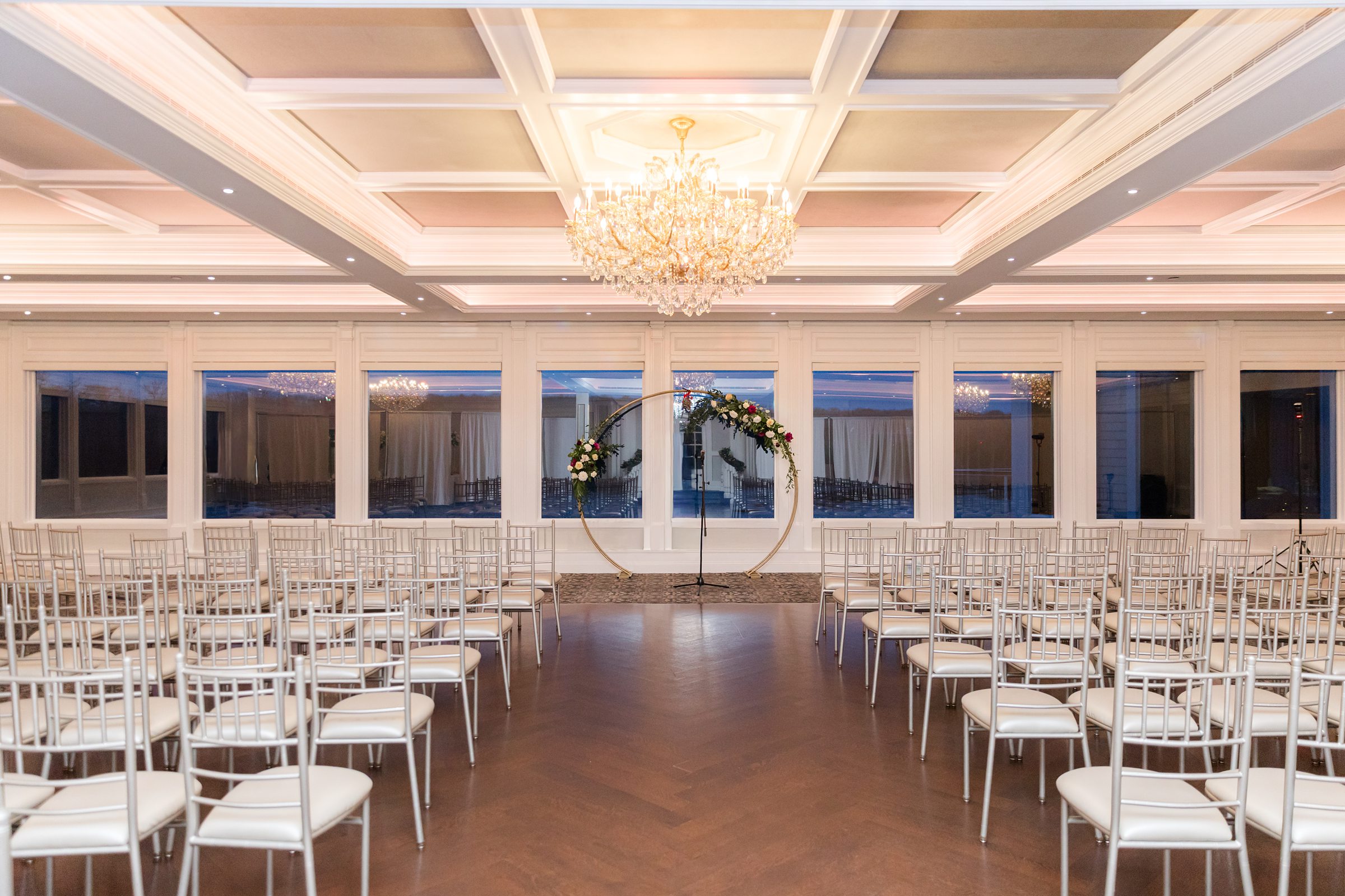 Elegant ceremony space with rows of white chairs leading to a circular floral arch, all set beneath a sparkling chandelier in a softly lit, romantic ballroom.