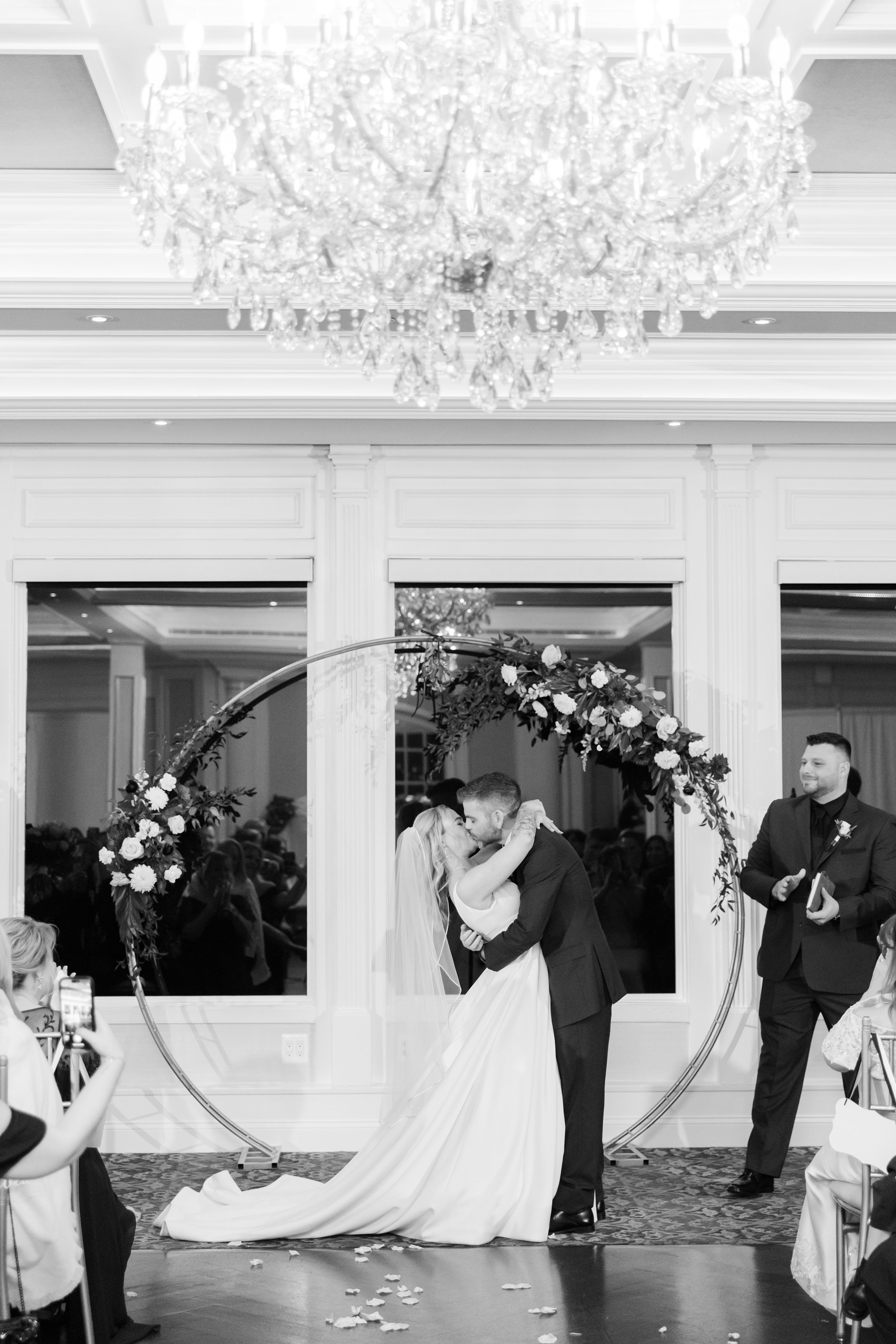 Bride and groom shares a tender kiss beneath a floral arch as guests look on, capturing a heartfelt and intimate ceremony moment.