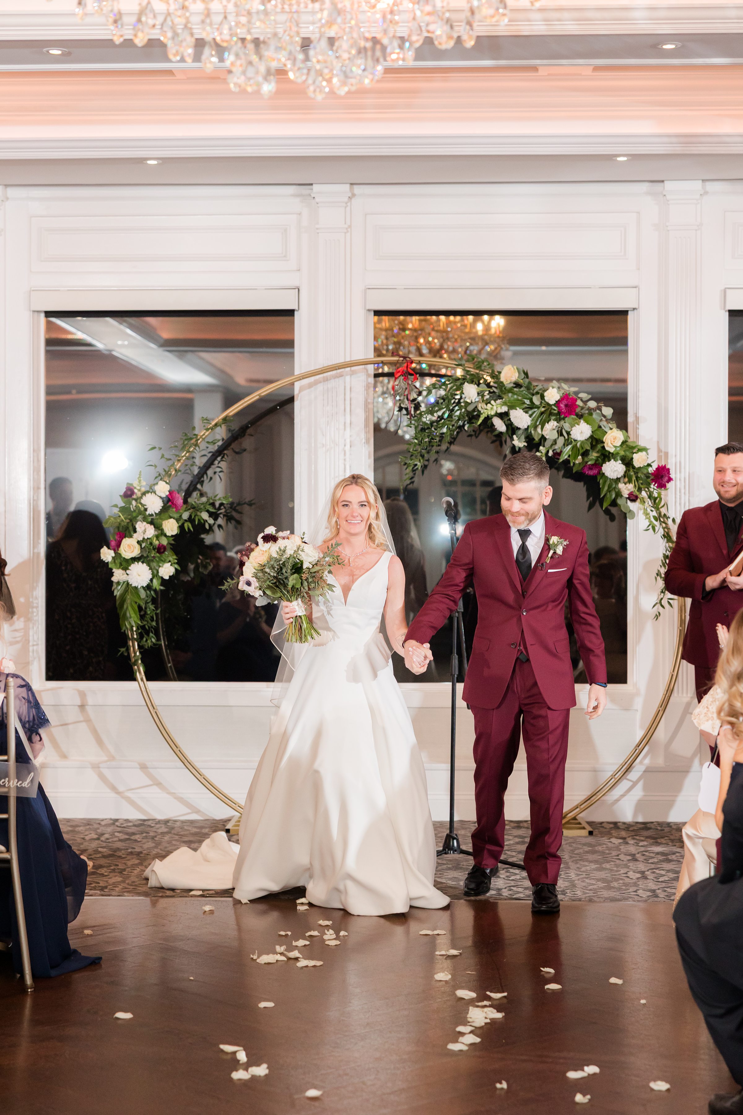 Bride and groom walk together under a circular floral arch, their expressions filled with happiness and anticipation.