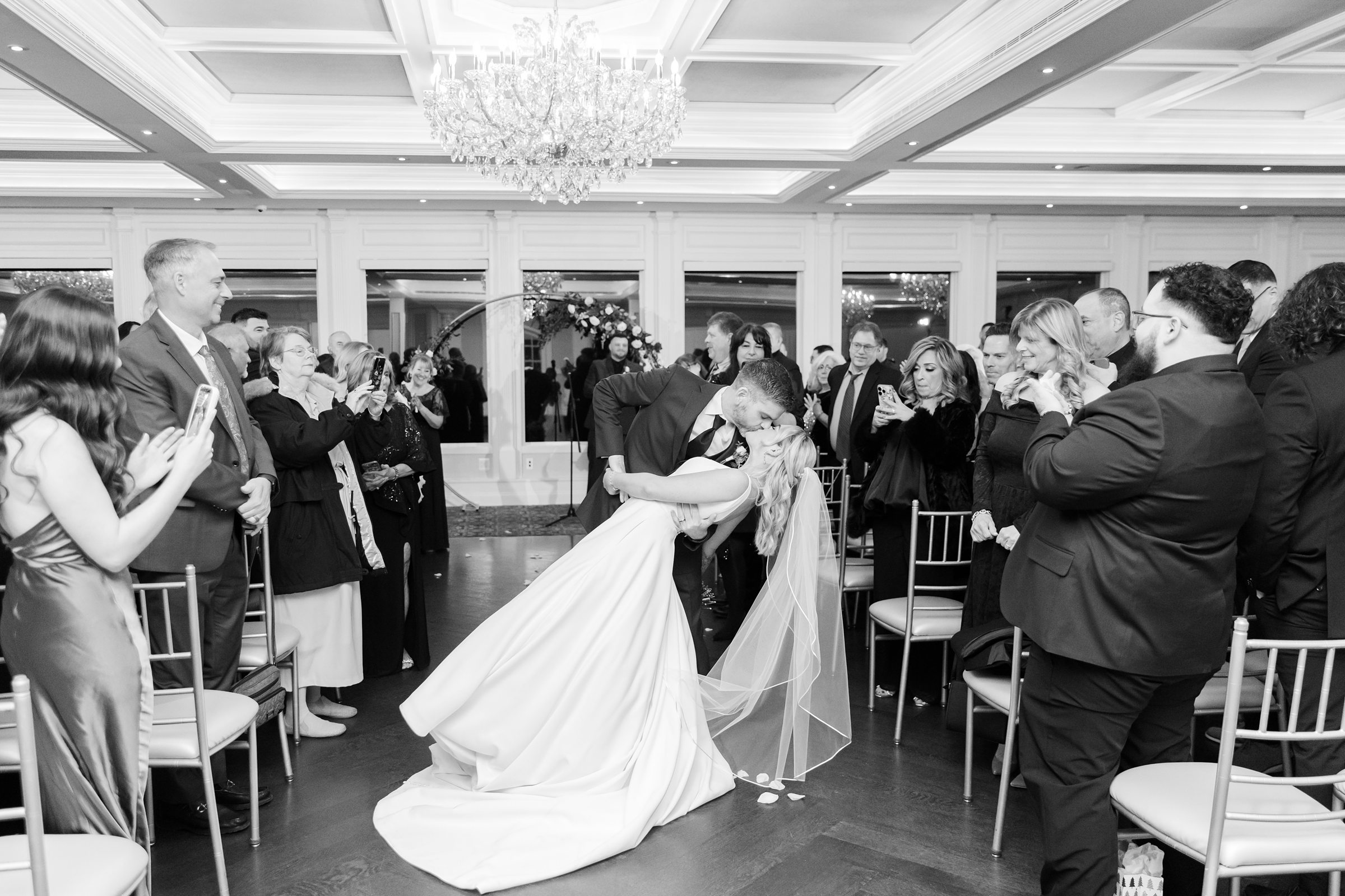 Bride and groom shares a dramatic dip and kiss beneath sparkling chandeliers, surrounded by loved ones in a joyful, cinematic moment.