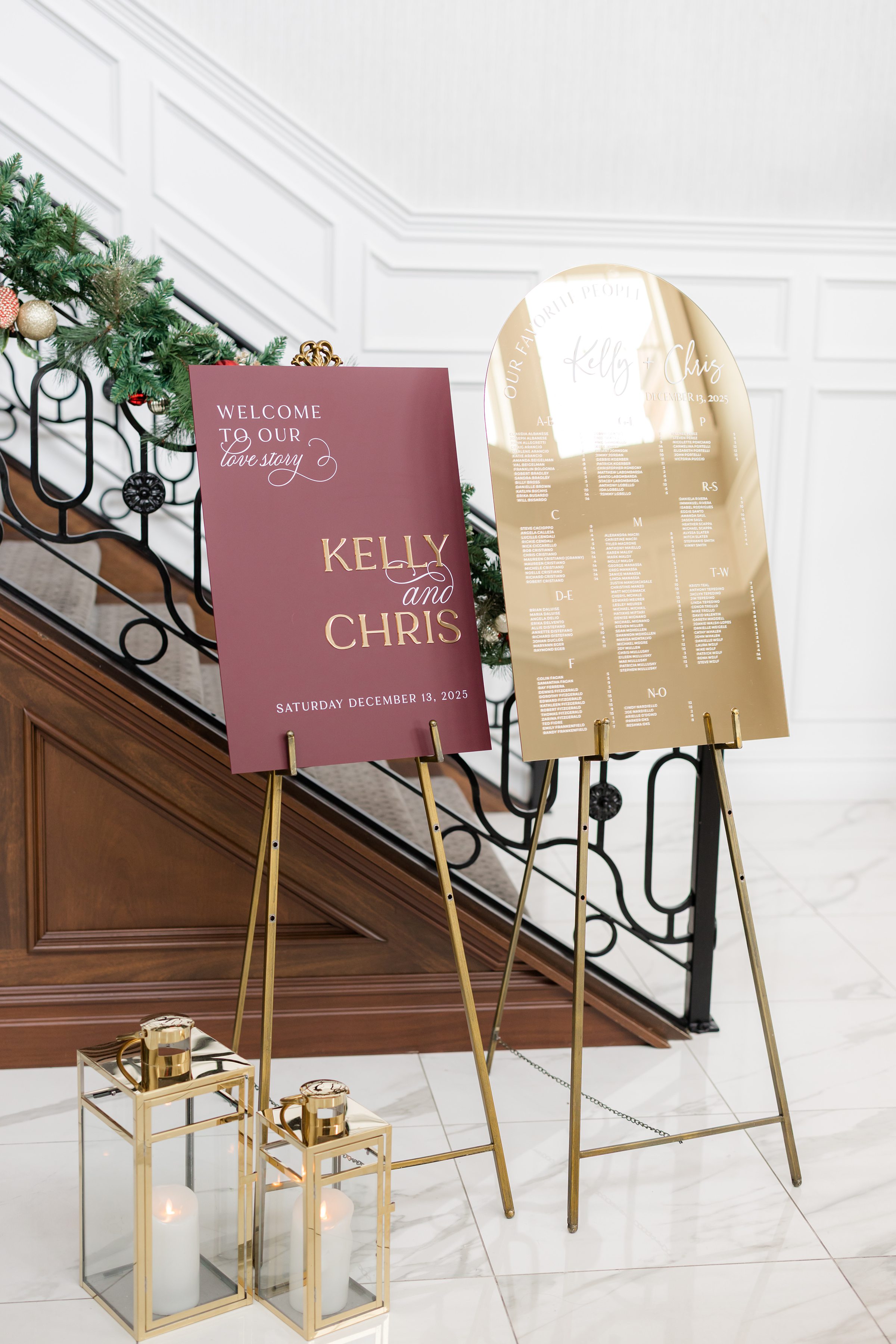 Wedding welcome display featuring burgundy and gold signage on easels, accented by candlelit lanterns and greenery.