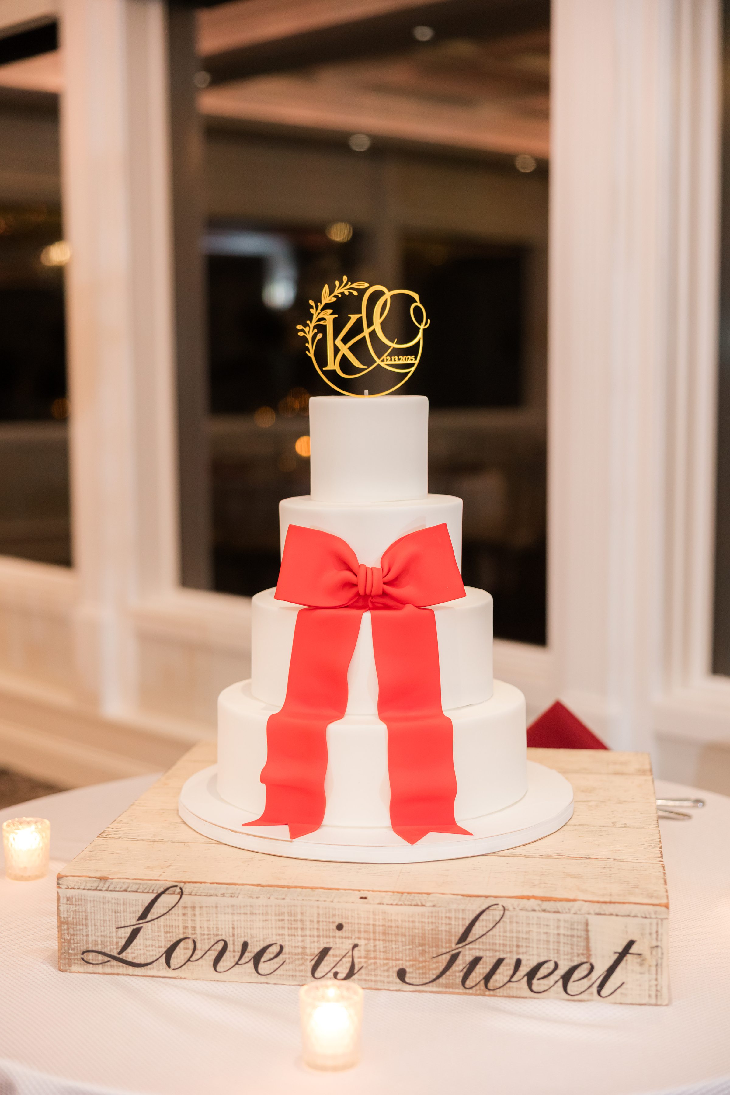White wedding cake adorned with a bold red bow and a gold monogram topper, displayed on a wooden stand in a softly lit reception setting.