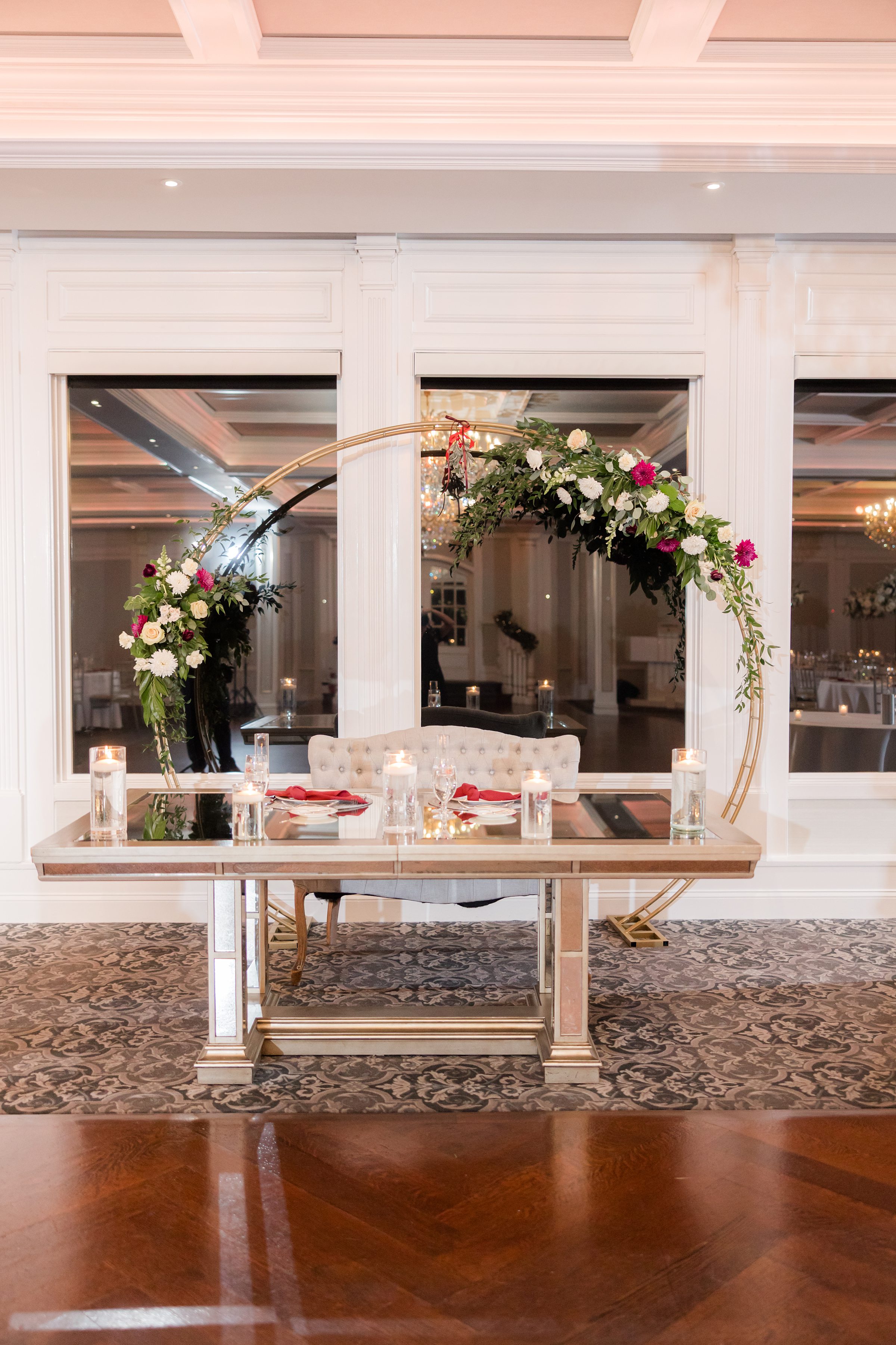 Elegant sweetheart table with candles and red accents, framed by a gold circular floral arch with white and blush flowers in a softly lit reception space.