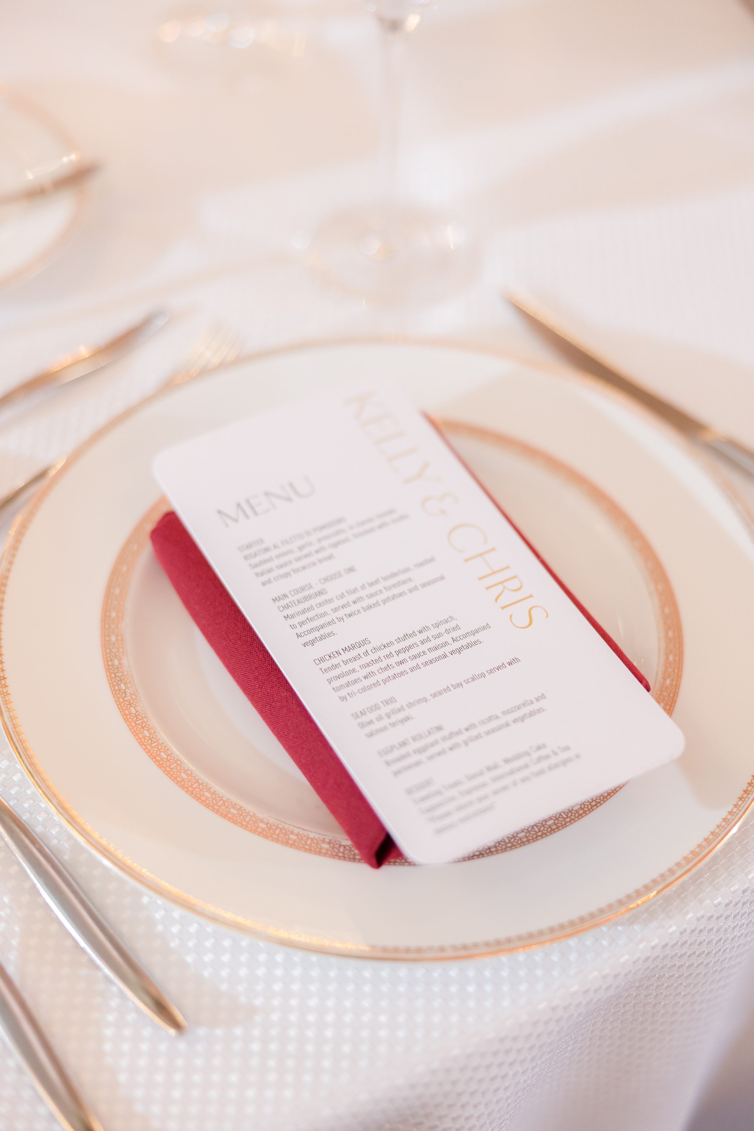 Elegant wedding place setting with a personalized menu card resting on a red napkin atop gold rimmed arranged on a softly lit reception table.