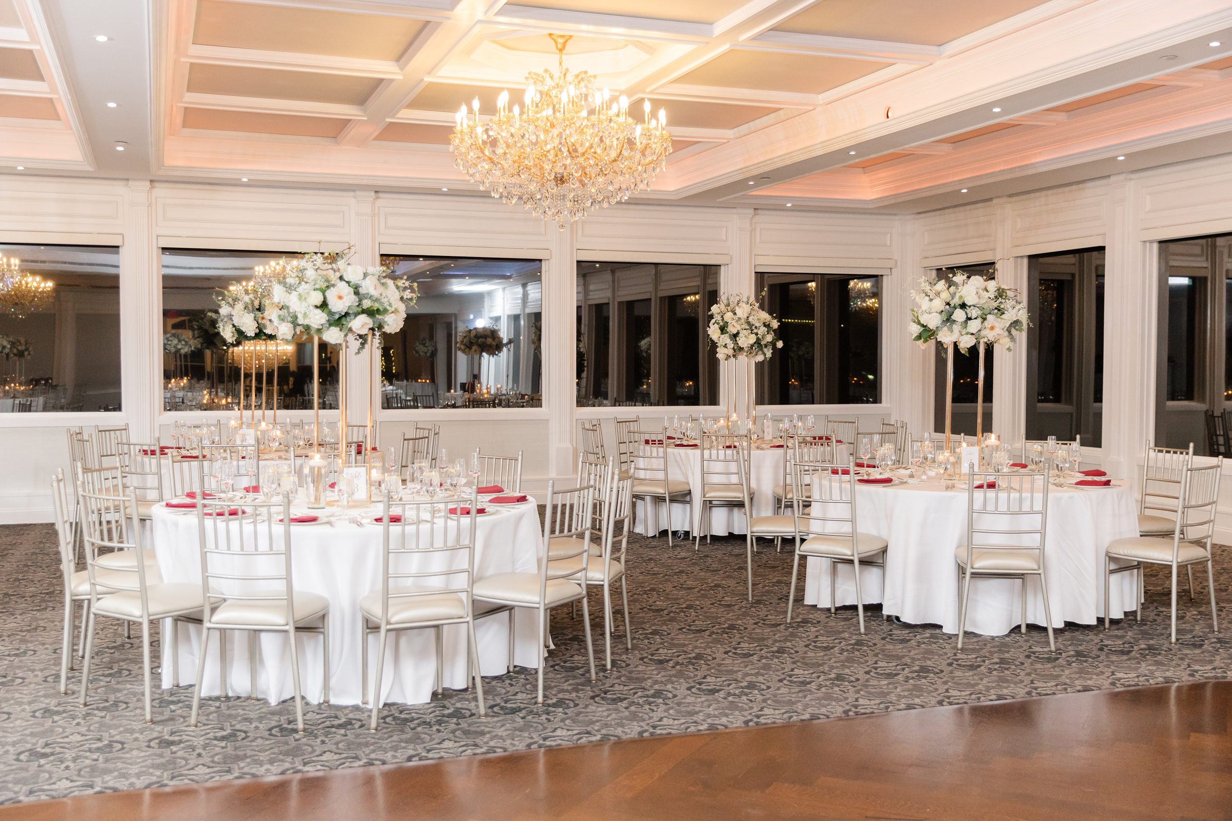 Elegant wedding reception ballroom with round tables, gold chairs, tall white floral centerpieces, and a crystal chandelier casting warm, romantic light.