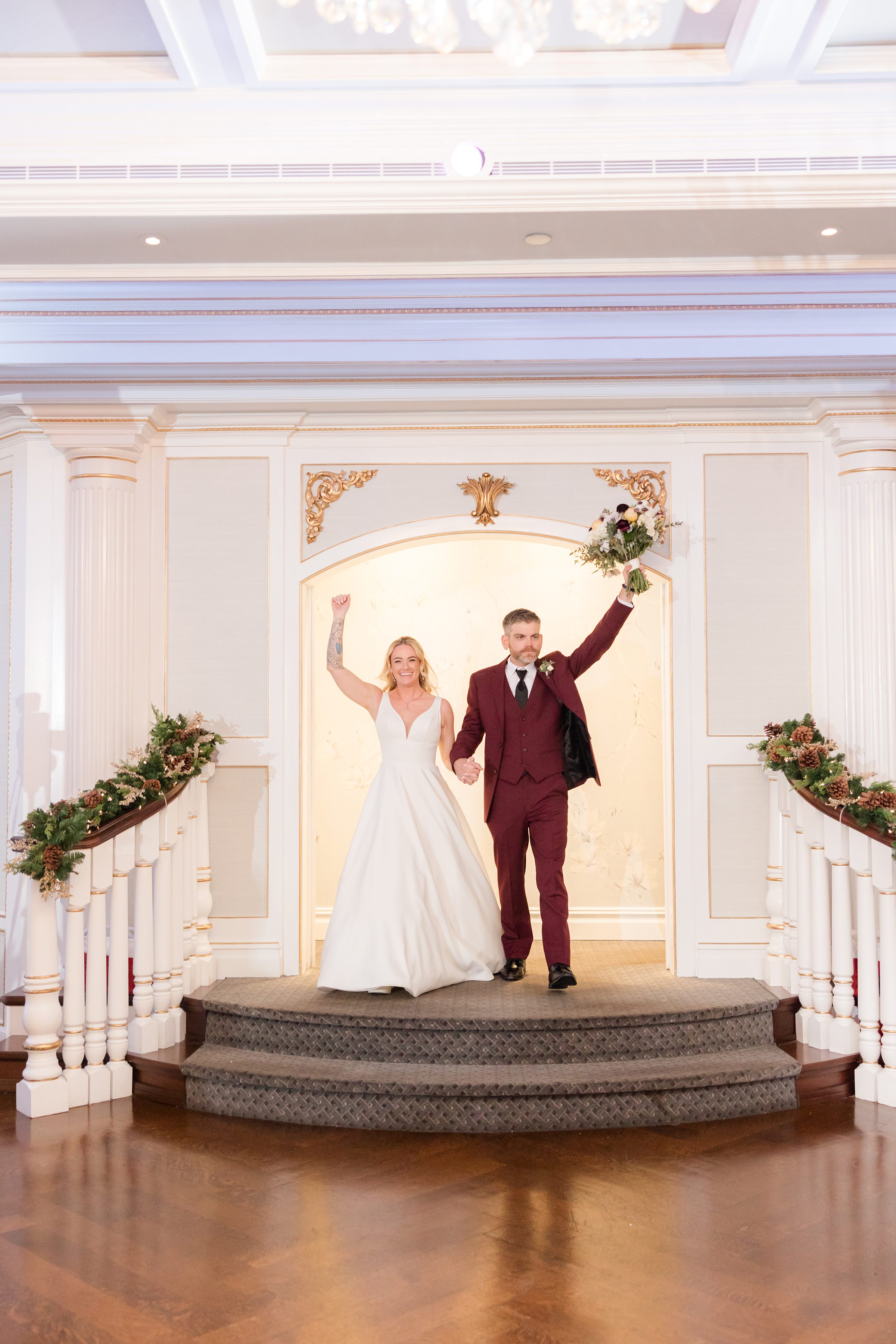Bride and groom making a joyful grand entrance, holding hands with arms raised, as they step into an elegant reception space decorated with greenery and warm lighting.