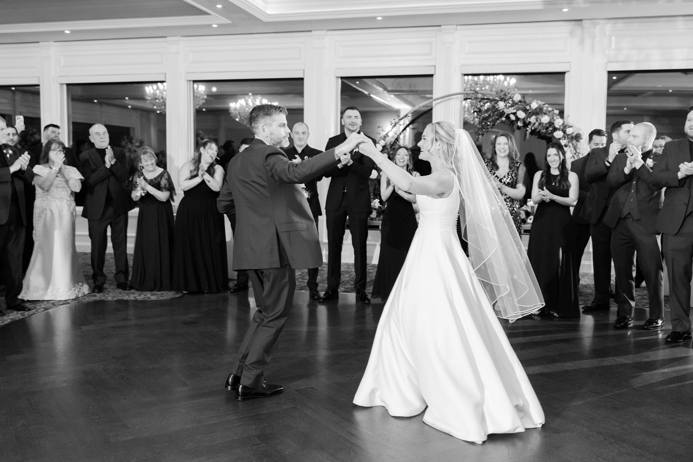 Bride and groom dancing together during their first dance, surrounded by smiling guests in an elegant ballroom.