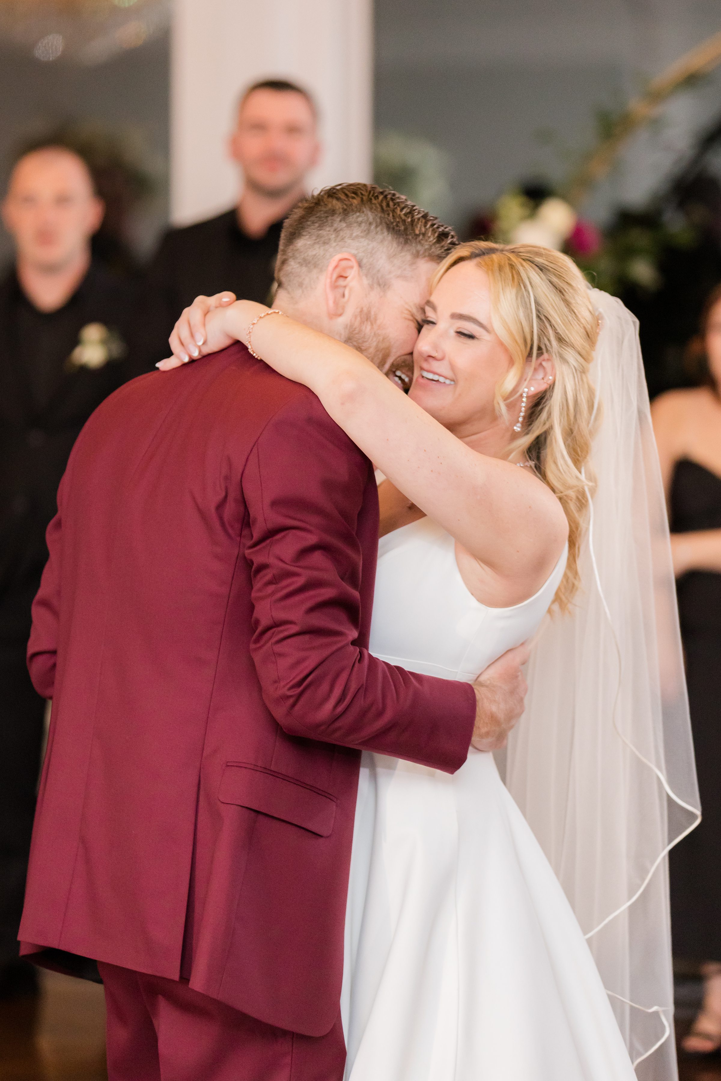 Bride and groom sharing a tender embrace during their first dance, smiling closely in a softly lit ballroom surrounded by guests.