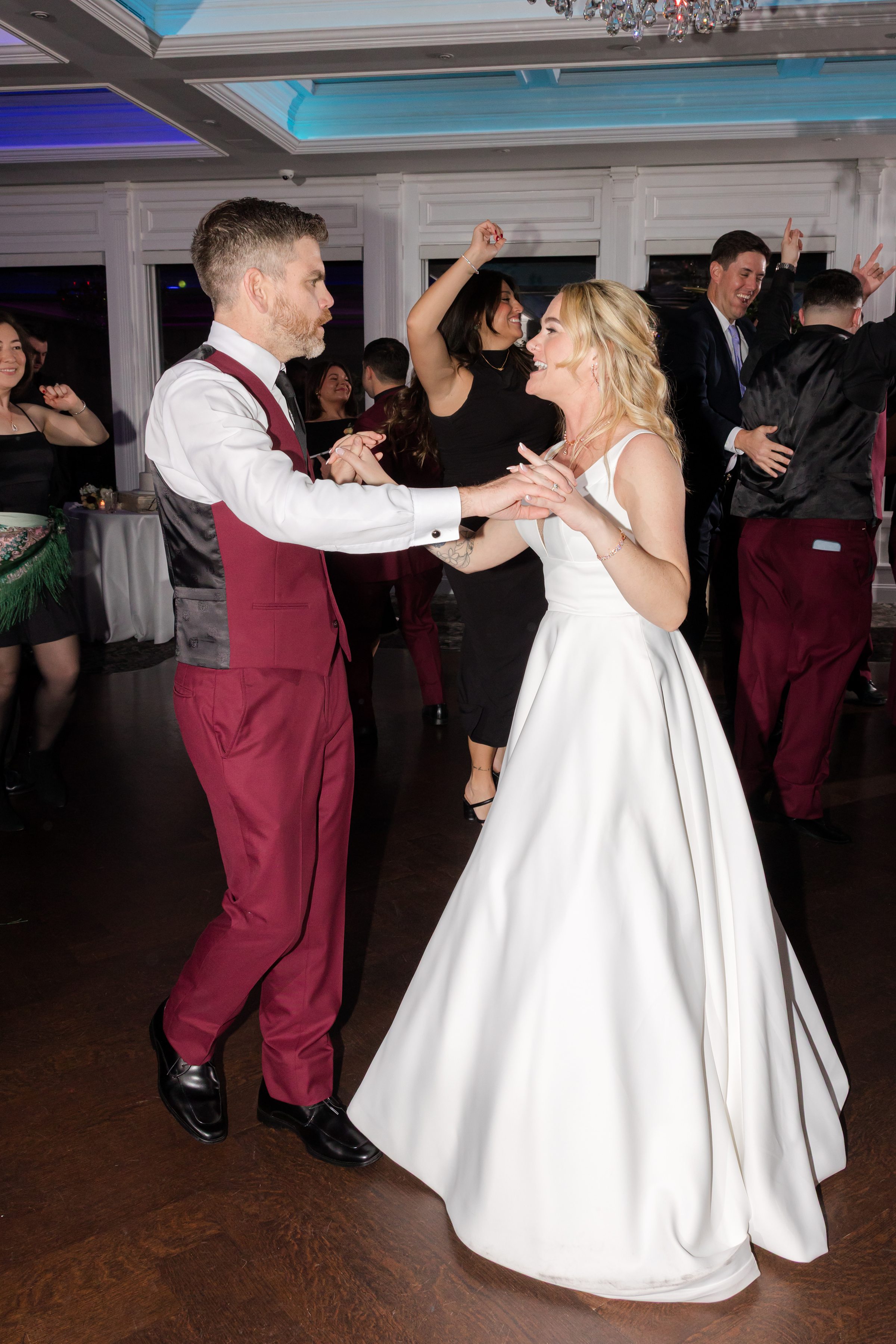Bride and groom dancing together on the reception dance floor, smiling and holding hands as guests celebrate around them under colorful lights.