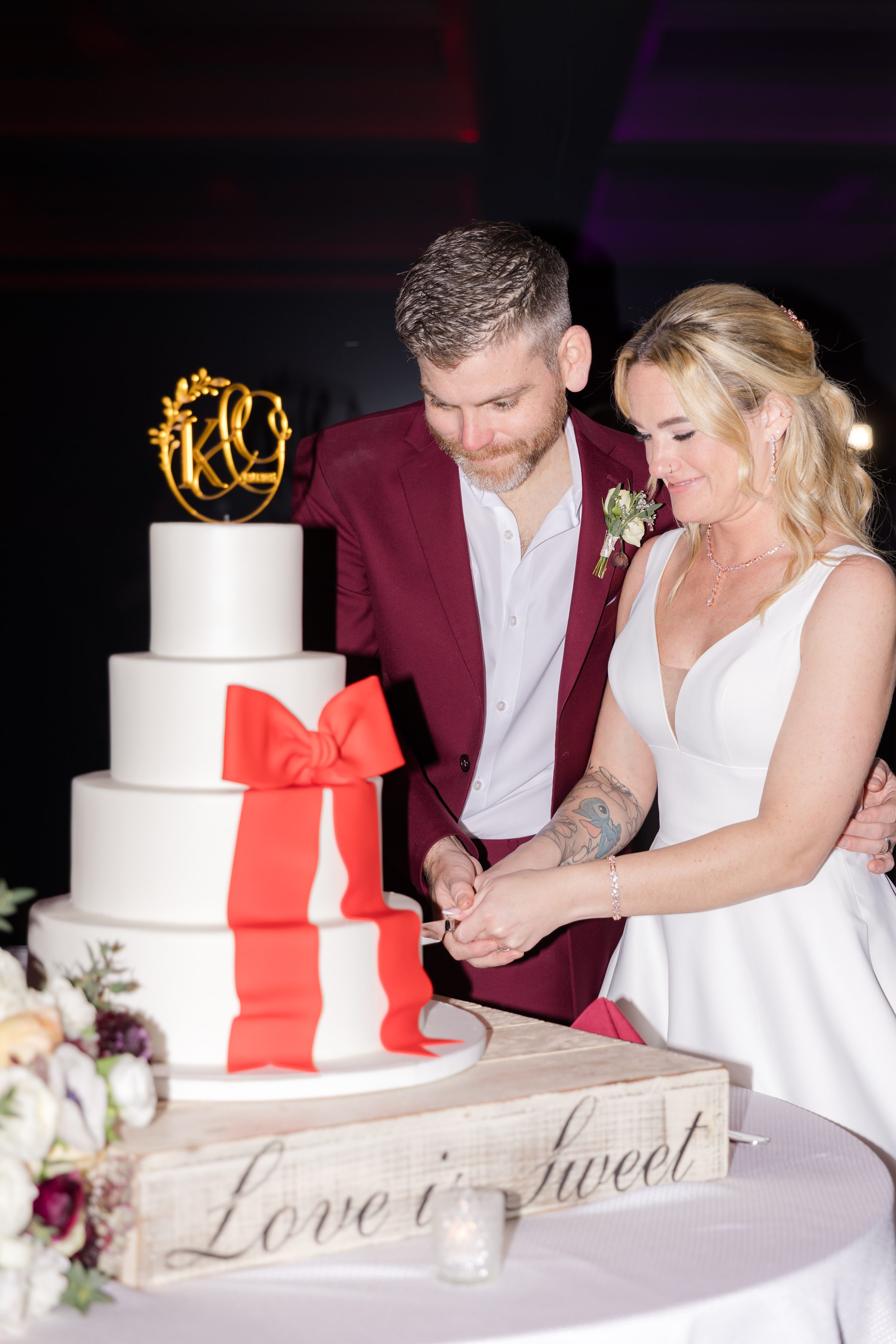 Bride and groom cutting their wedding cake together, smiling closely beside a white tiered cake with a red bow and gold monogram topper.