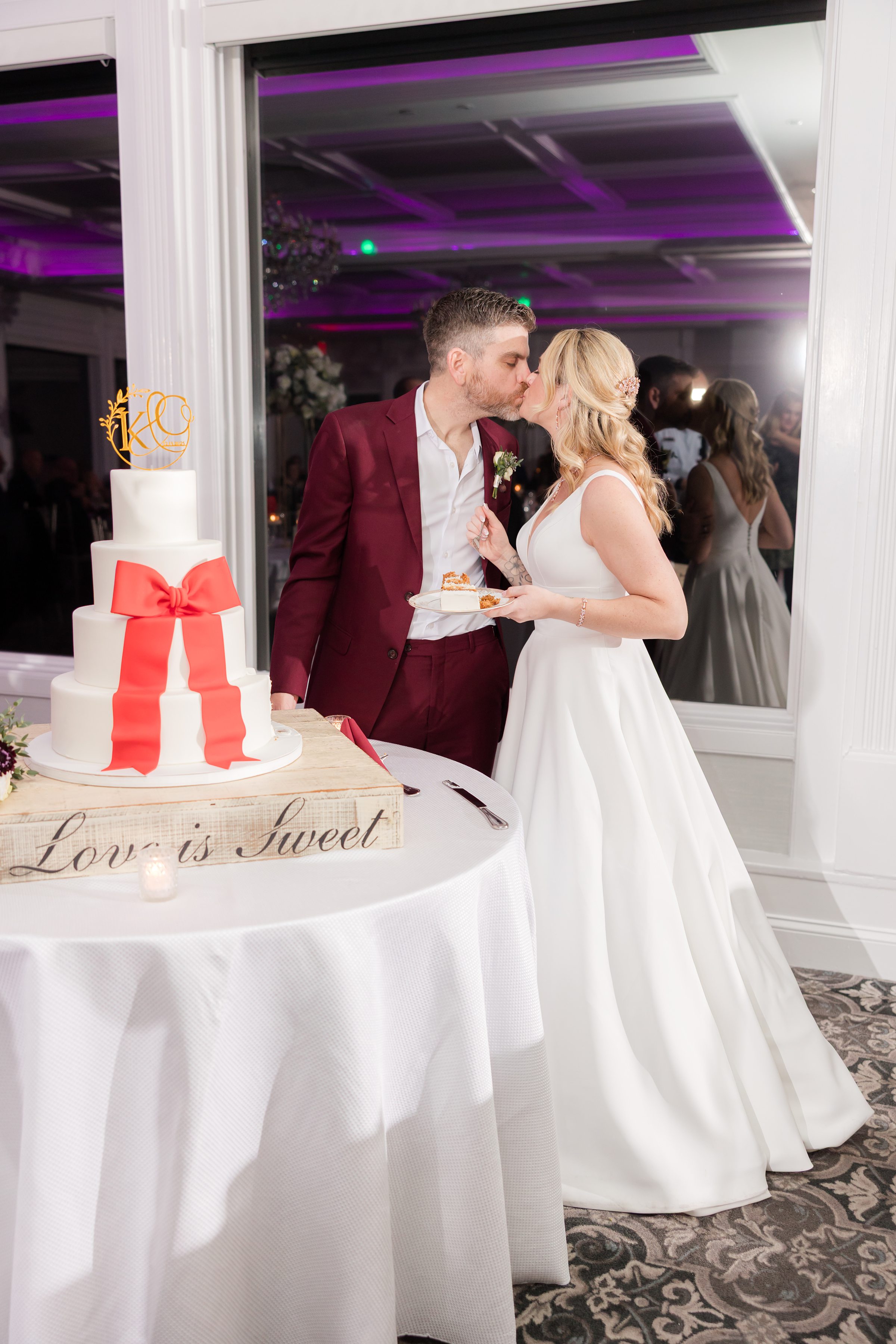 Bride and groom sharing a kiss beside their wedding cake, holding a slice on a plate next to a white tiered cake with a red bow in a softly lit reception.