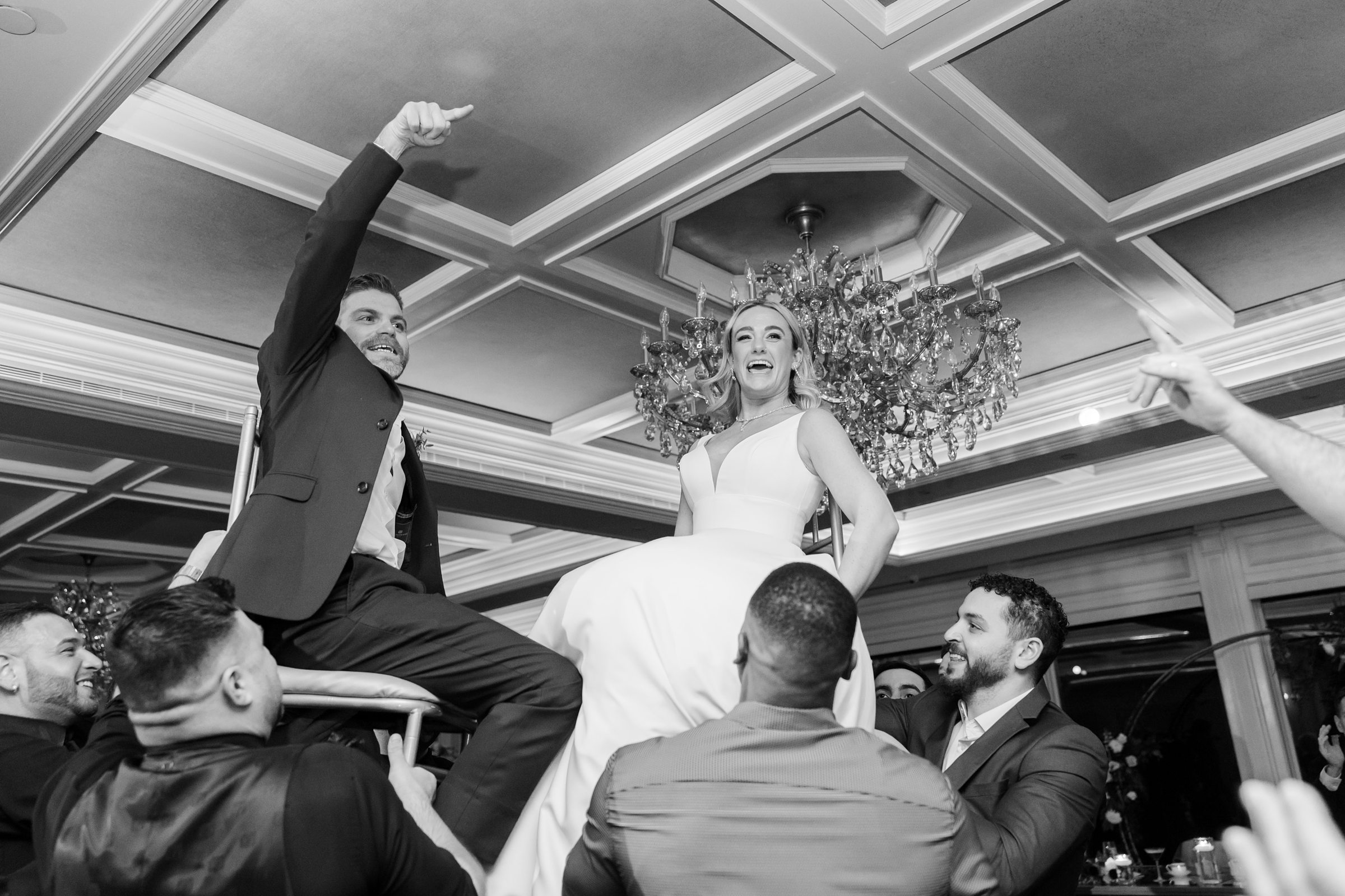 Bride and groom lifted on chairs during a lively reception celebration, smiling and cheering beneath a grand chandelier.