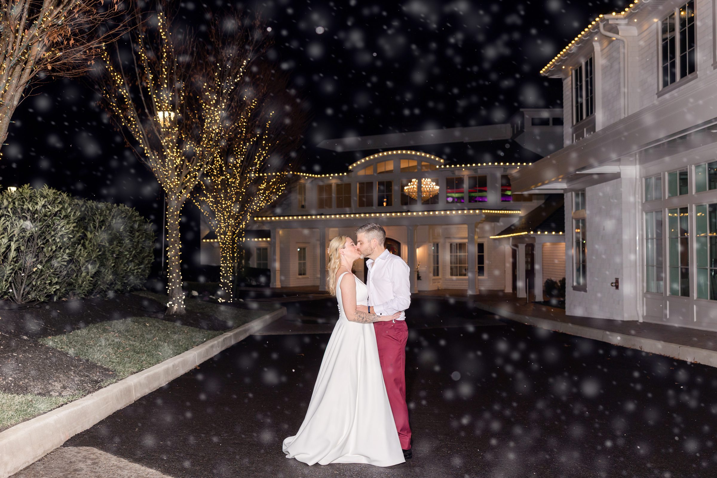 Bride and groom sharing a romantic kiss in falling snow outside a beautifully lit venue wrapped in twinkling lights at night.