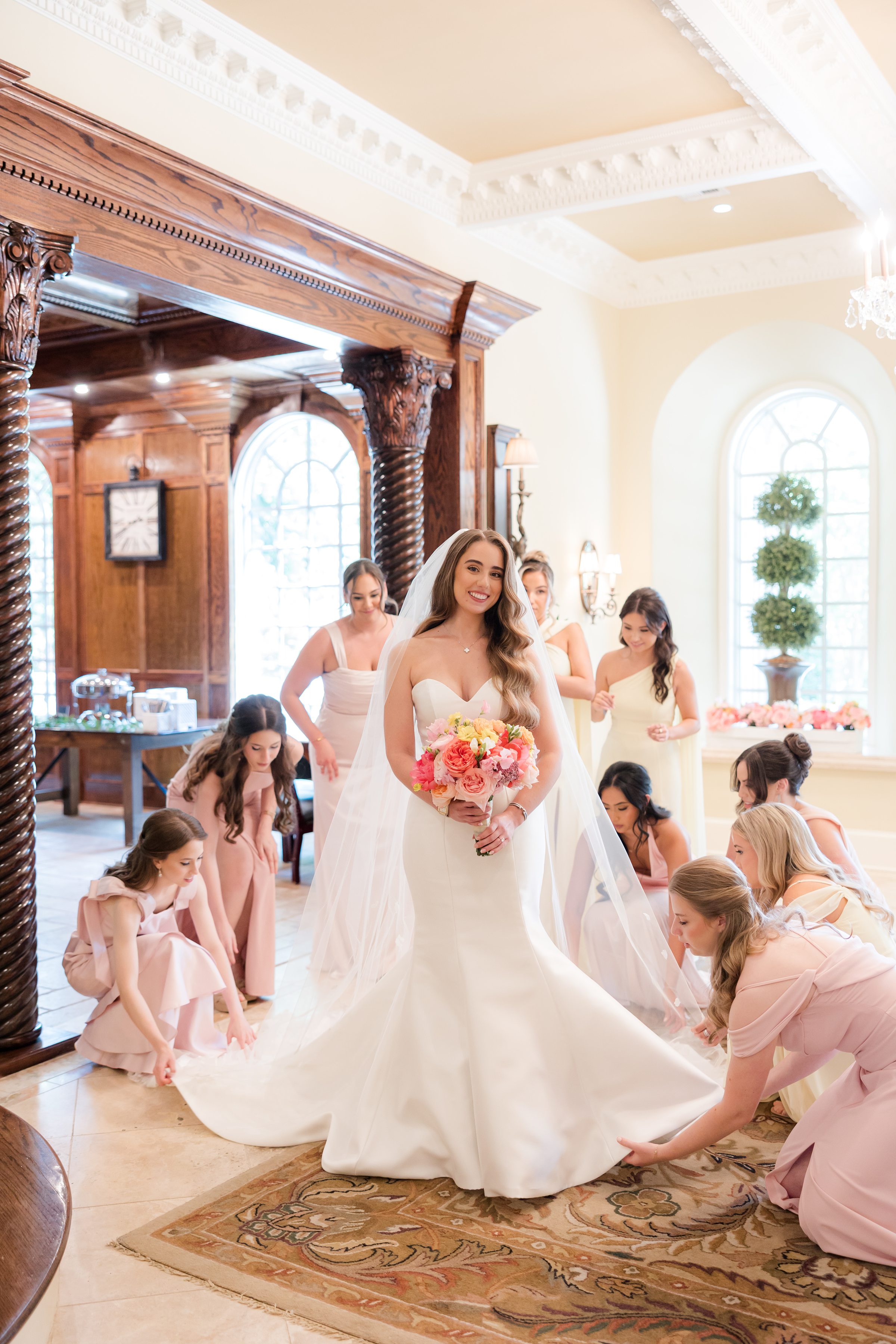 Bride smiling at the camera while her bridesmaids in pink and cream dresses fix her train