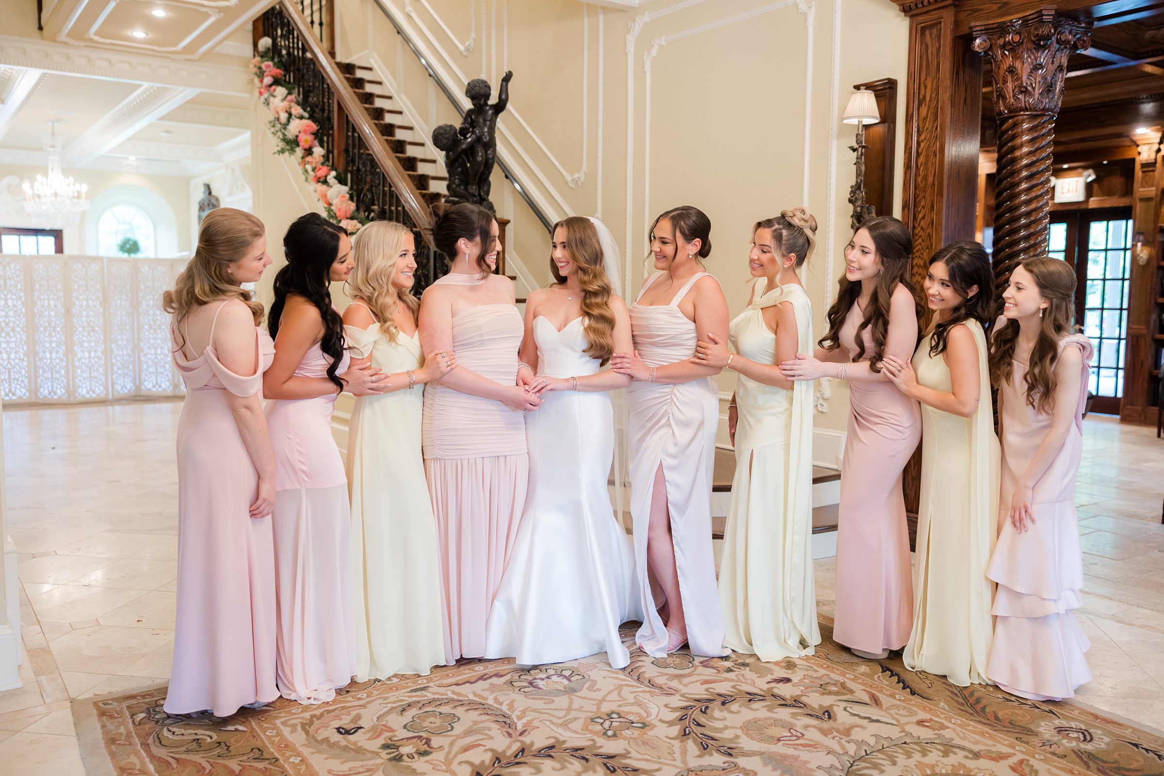 Bride talking with bridesmaids under the stairs. Bridal party is wearing pink, yellow, and cream dresses.