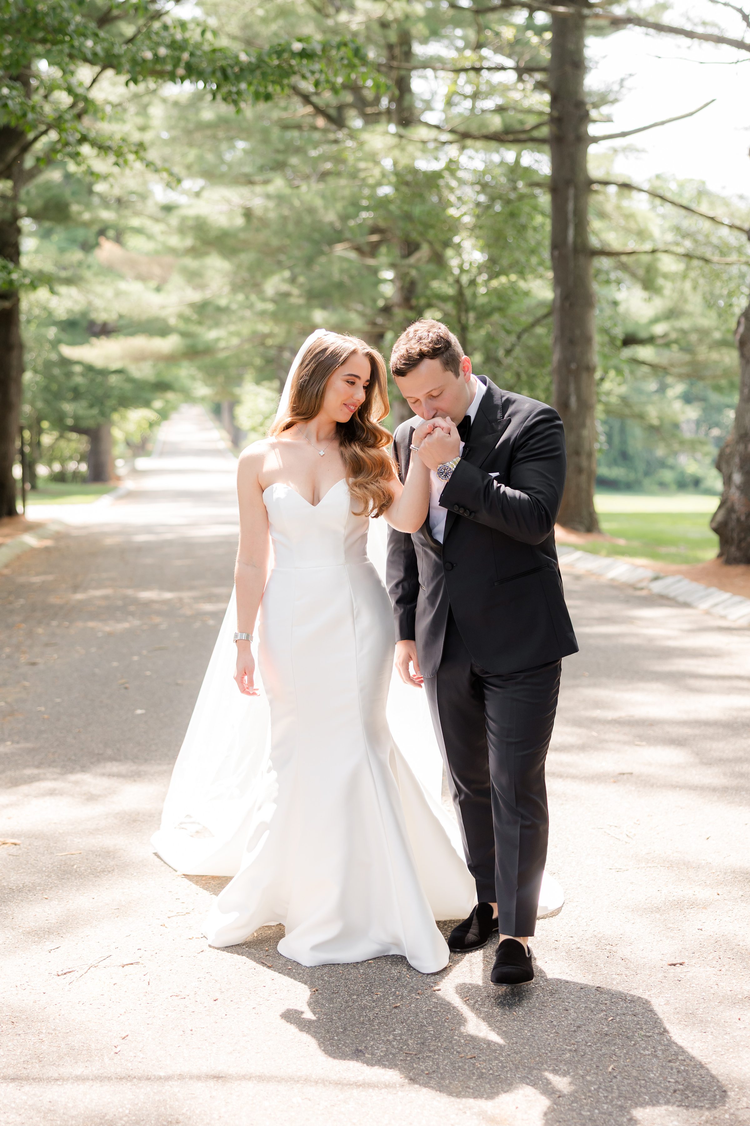 Full-body portrait of groom holding and kissing the bride's hand, along a road surrounded by trees and grassland