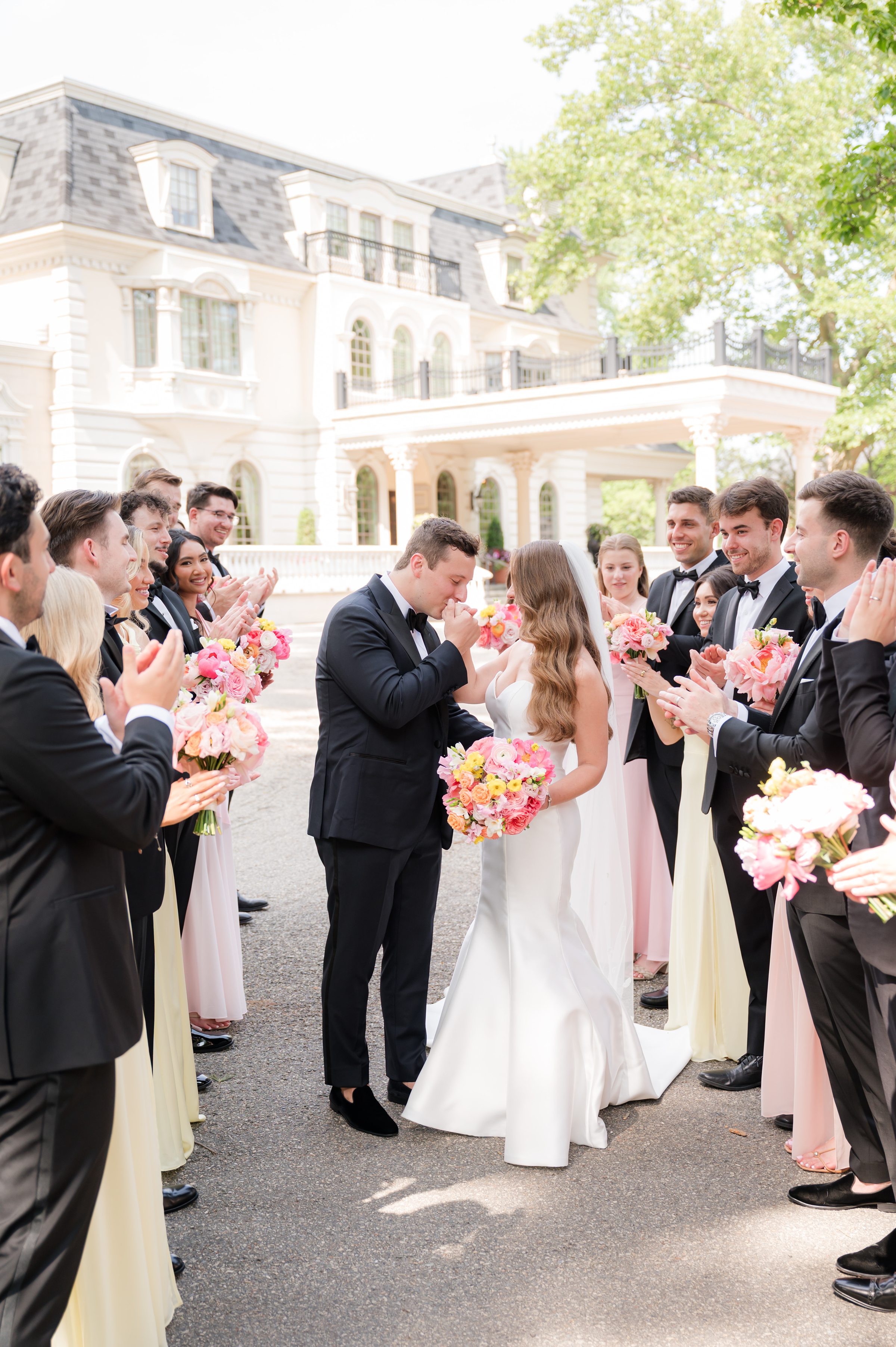 Wedding party lined up side-by-side to let the couple pass as the groom holds the bride's right hand and kisses it, while the bride holds her bouquet with her left hand