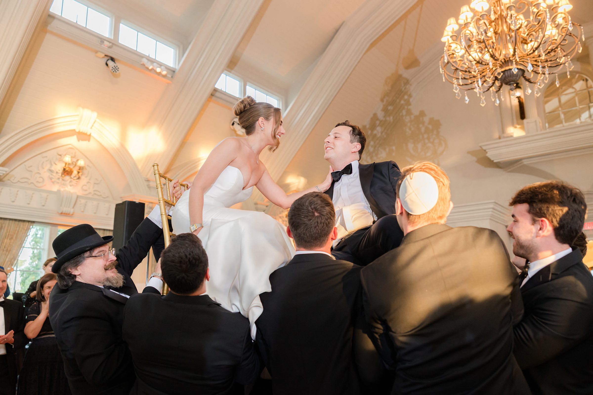 The bride and groom are being lifted by men guests, the bride is sitting on a chair