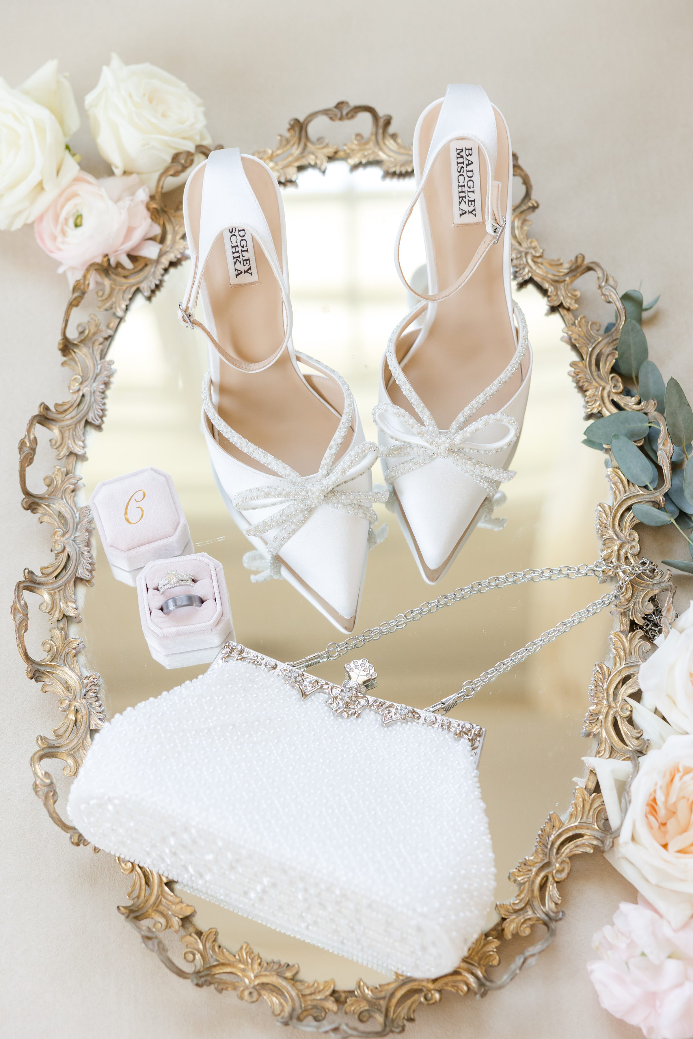 a detail shot of bride's wedding shoes, wedding ring, white purse placed on the golden mirror with cream and pink flowers on the sides 