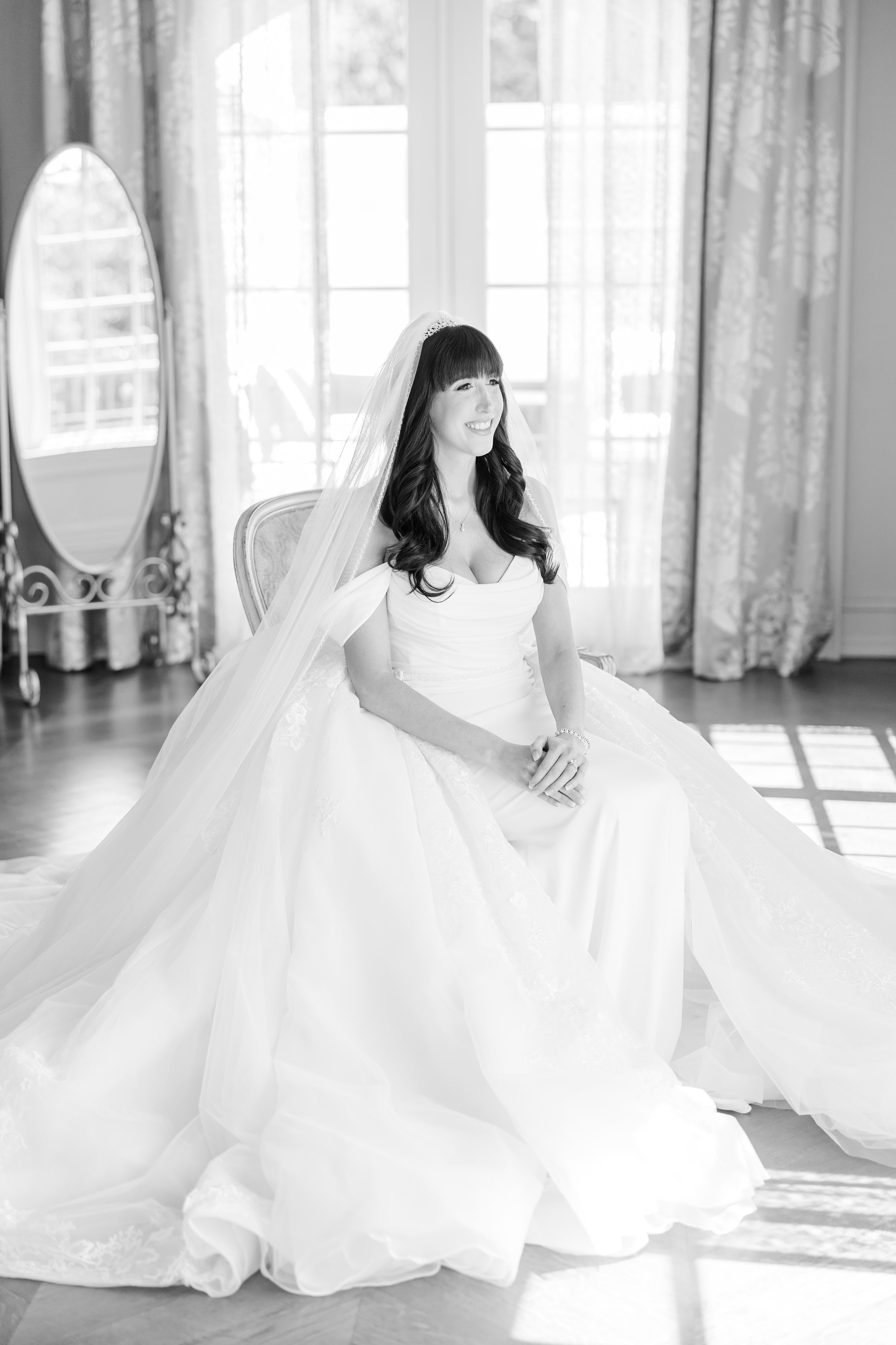 a solo photo of bride sitting, wearing her white off-shoulder wedding dress with long veil while smiling, photo is in black and white