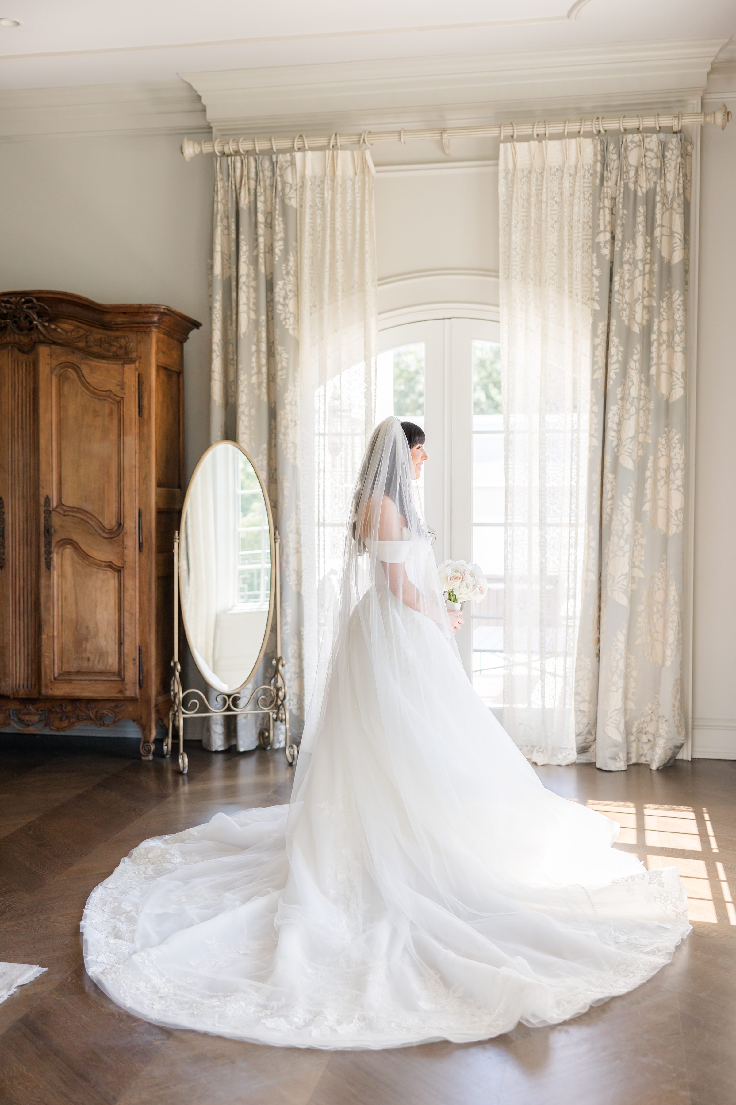 full portrait of back of the bride's dress wearing a long veil facing the window with mirror on the side