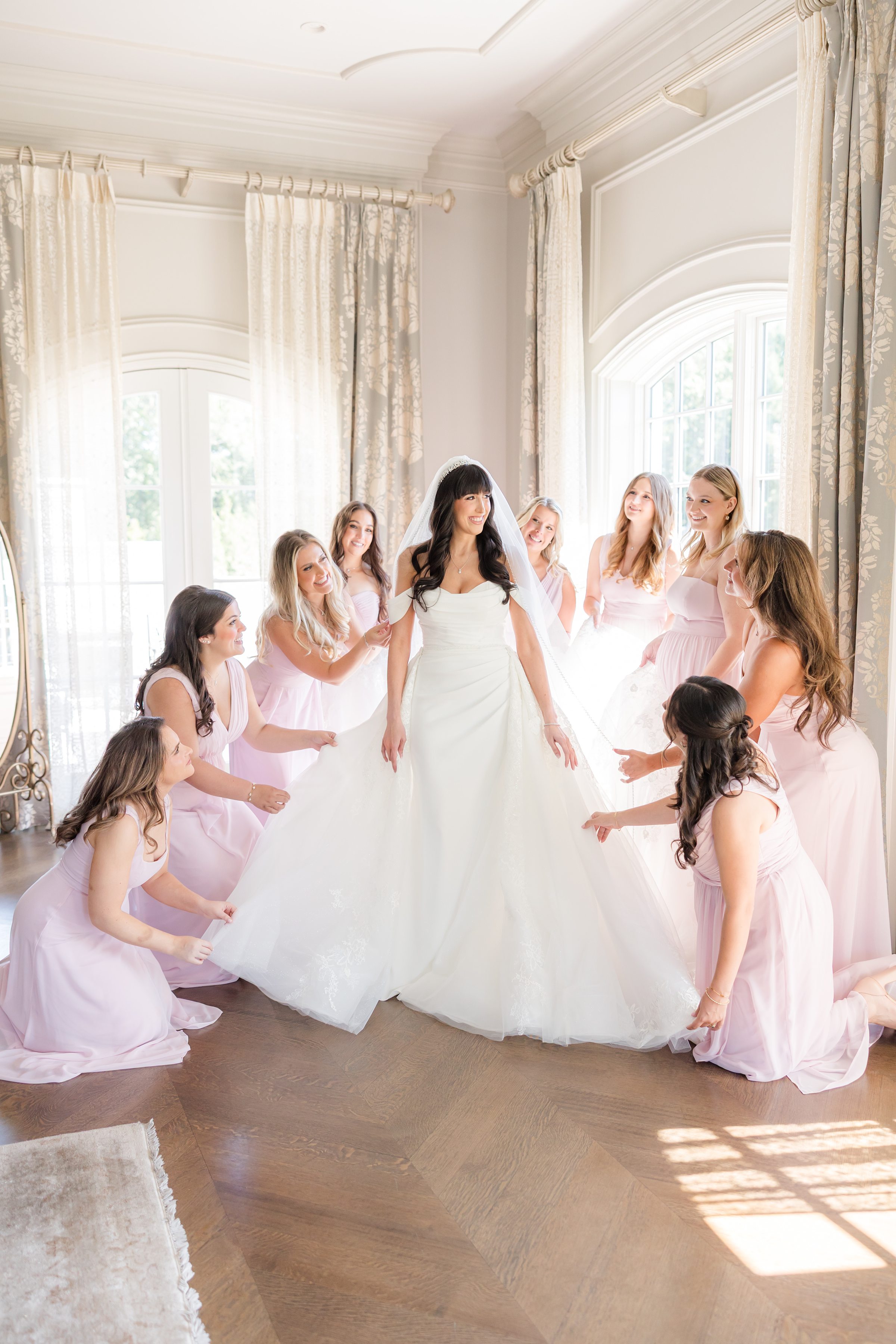 bride standing with bridesmaids while holding her wedding dress, smiling at each other 