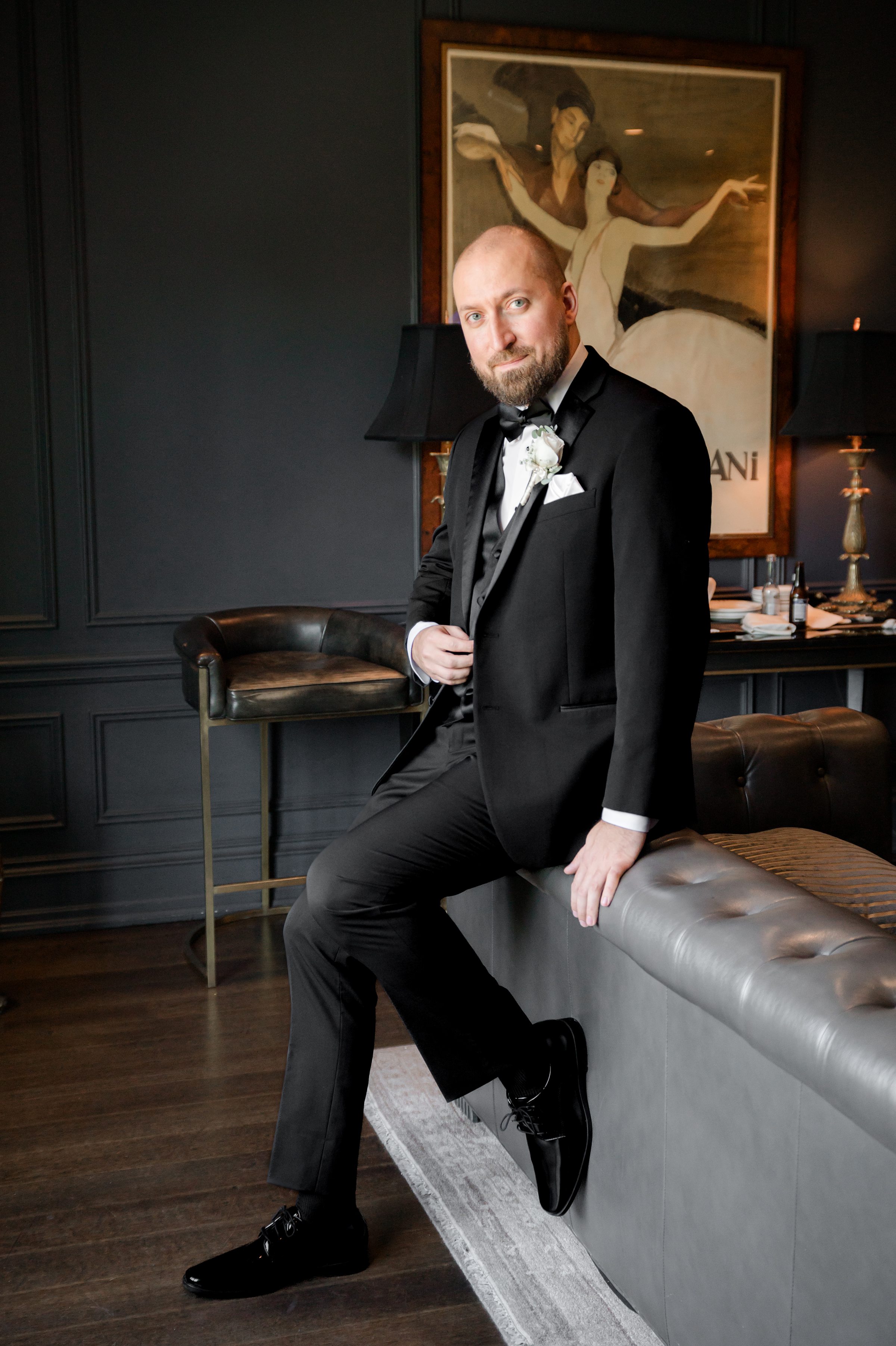 full body picture of groom sitting on the couch wearing black tuxedo with a painting in the back