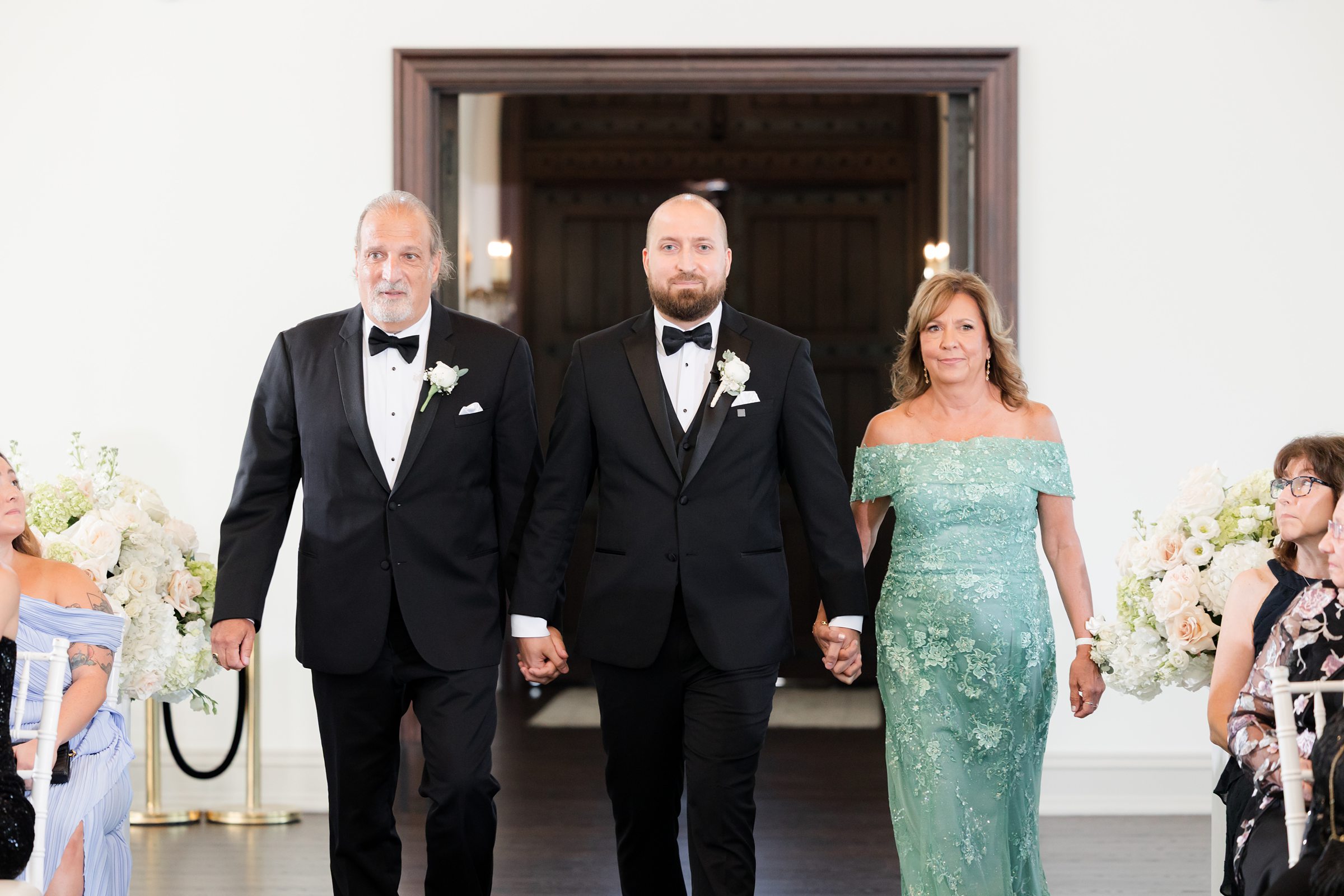 Groom and his parents walking him down the aisle while holding his hands. Mother of the groom, is wearing a teal, off-shoulder dress
