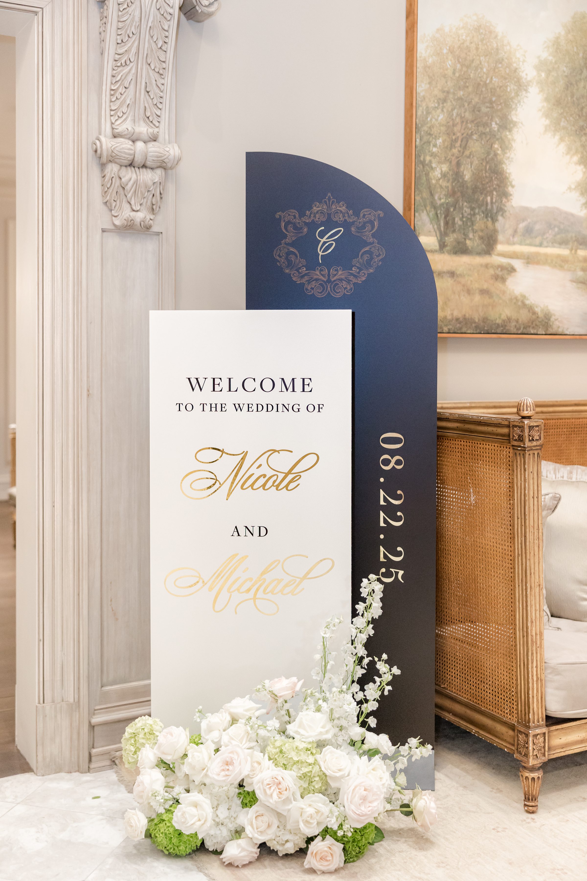 Welcome post in the reception hall of Park Chateau Estate, with white, gold, and navy blue color scheme, and flowers at the bottom. Written on the board is "WELCOME TO THE WEDDING OF Nicole and Michael 08.22.25"