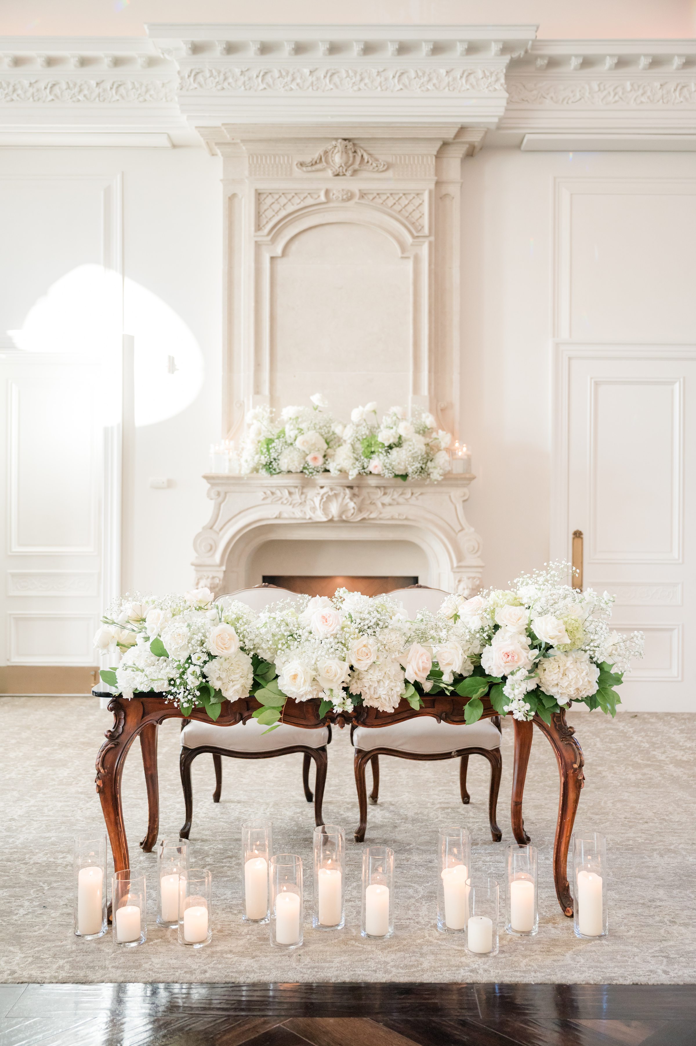 Photo of sweetheart table at Park Chateau Estate's ballroom, focusing on white flowers on top of the table and fireplace, with candles at the table's feet