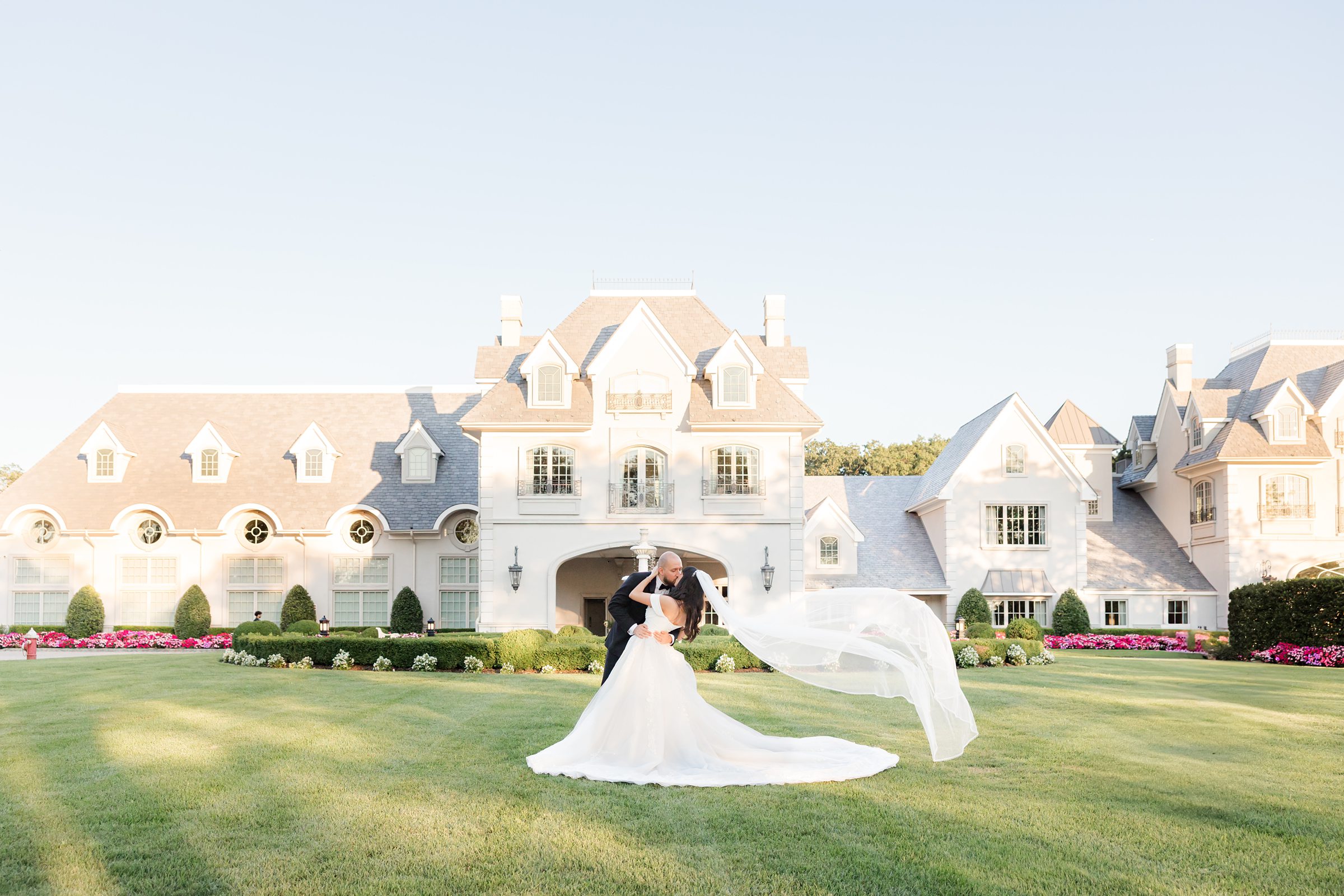Landscape photo of the bride and groom at Park Chateau Estate's facade. Groom is leaning into the bride, supporting her back, while she bends down holding his head, and her veil blown by the wind