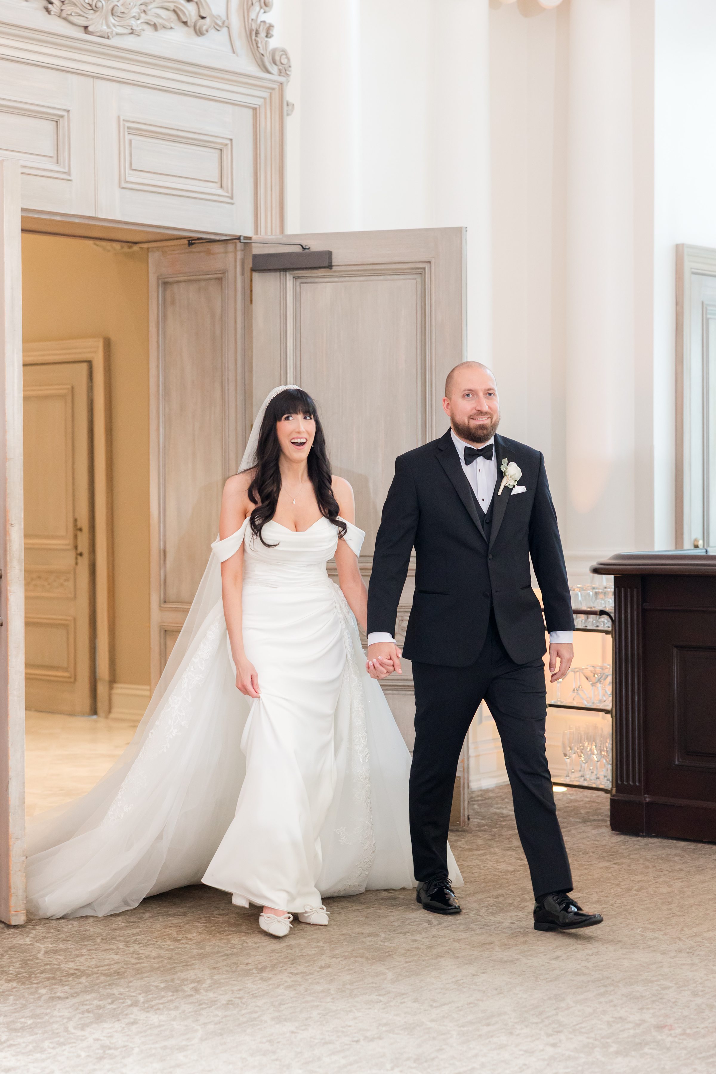 Bride and groom walking into the ballroom hand-in-hand for the first time. Groom keeps a subtle smile while the bride is pleasantly surprised with her mouth open