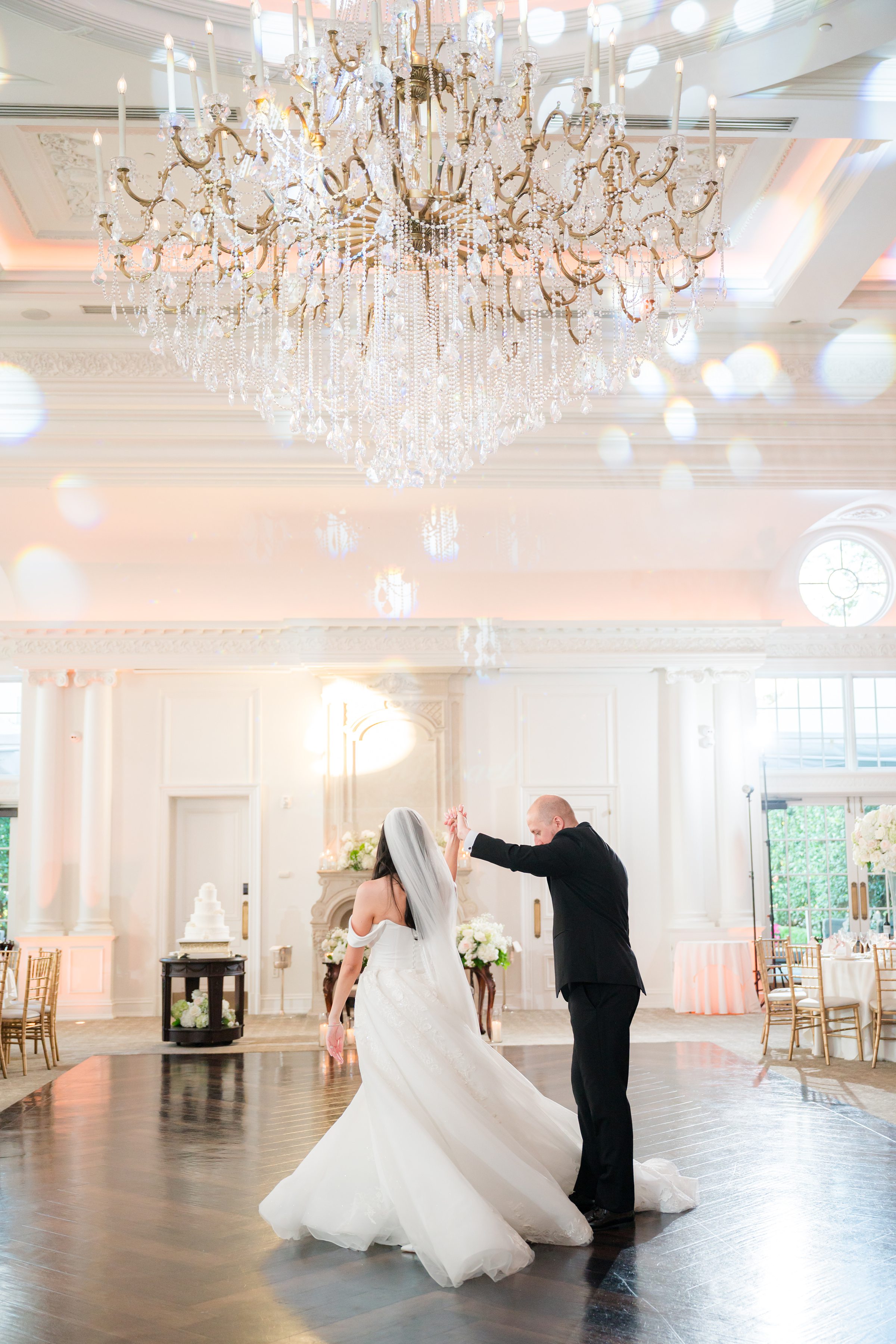Photo of bride and groom rehearsing their first dance in an empty ballroom with a big chandelier on top. Groom is helping the bride do a spin