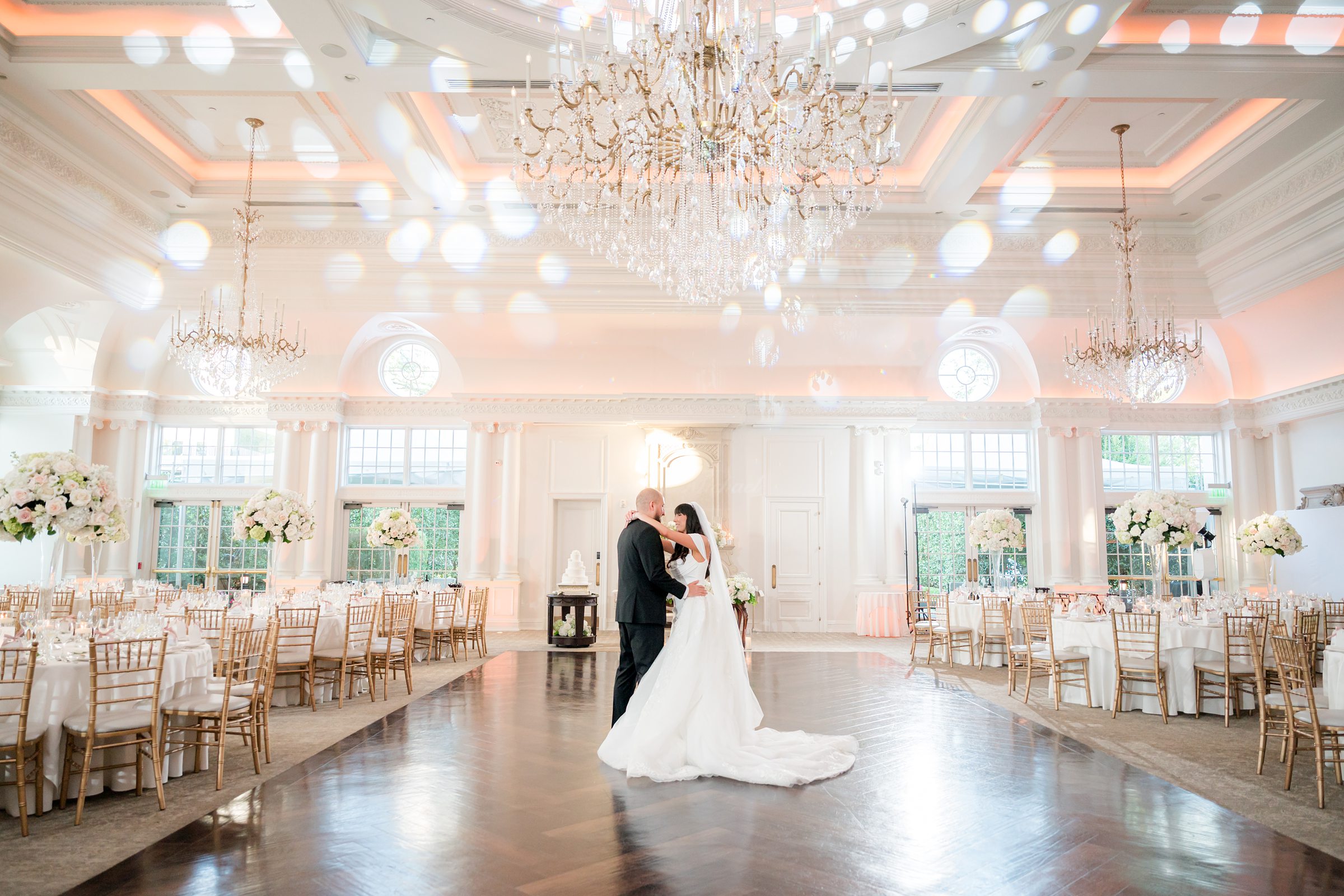 Bride and groom dancing on a wooden dancefloor, in an empty ballroom with a big chandelier on top. Groom is holding the bride's waist while her hand is on his shoulders