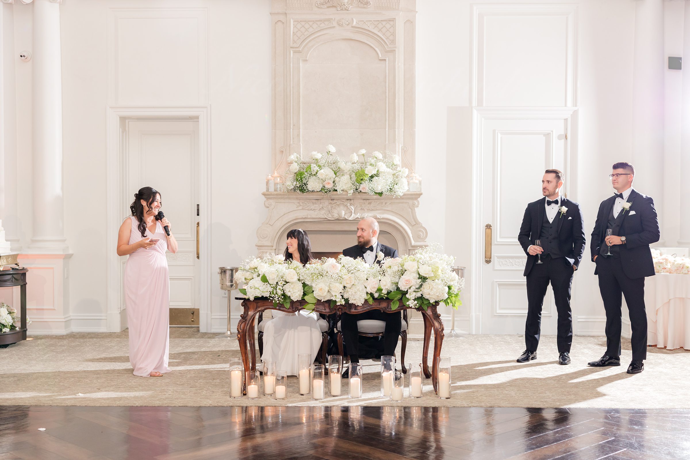 Bride and groom looking to their right while the bridesmaid is delivering her speech. To the left are 2 groomsmen holding a bottle of champagne