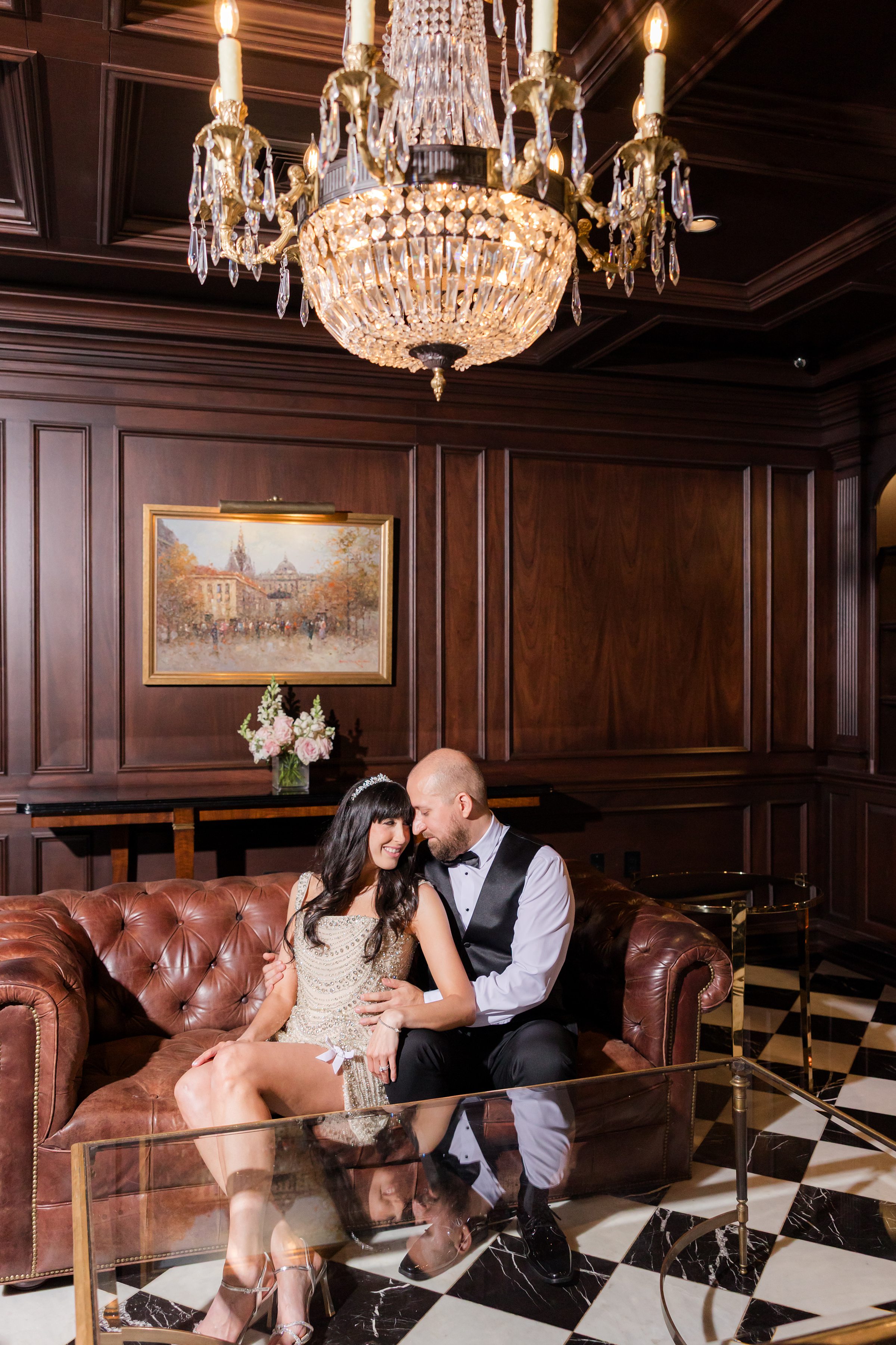 Bride and groom sitting in a brown sofa in a wooden room, with a bright warm chandelier on top