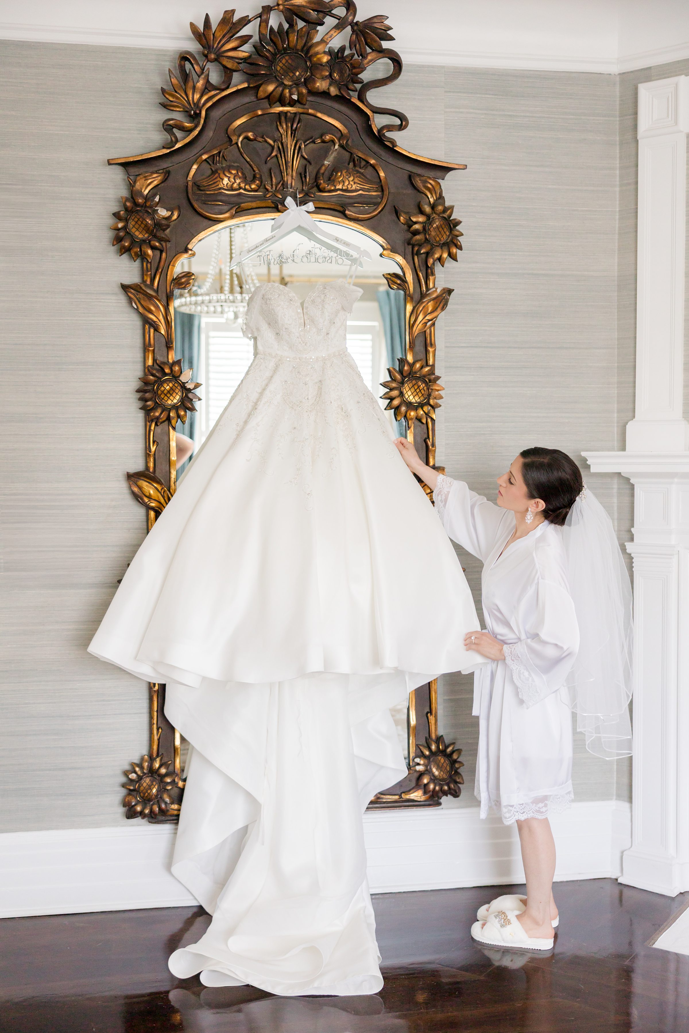 Bride wearing a white robe and a short veil, looking at her wedding dress hung on a brown and brass mirror with sunflower accents, while holding the bottom of the dress