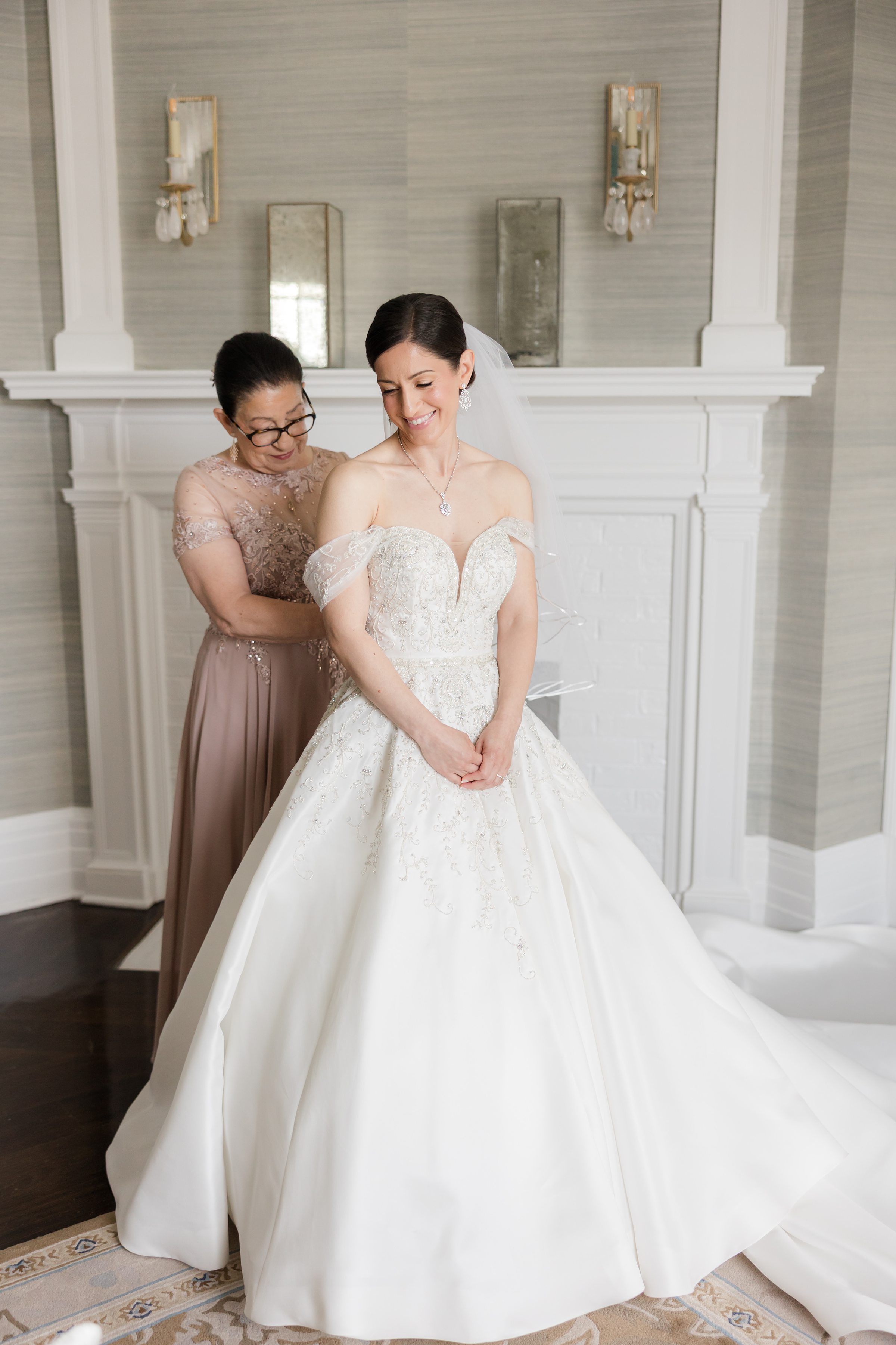 Mother of the bride fixing the back of her dress. Bride is wearing an off-shoulder ballgown style dress with a sweetheart neckline