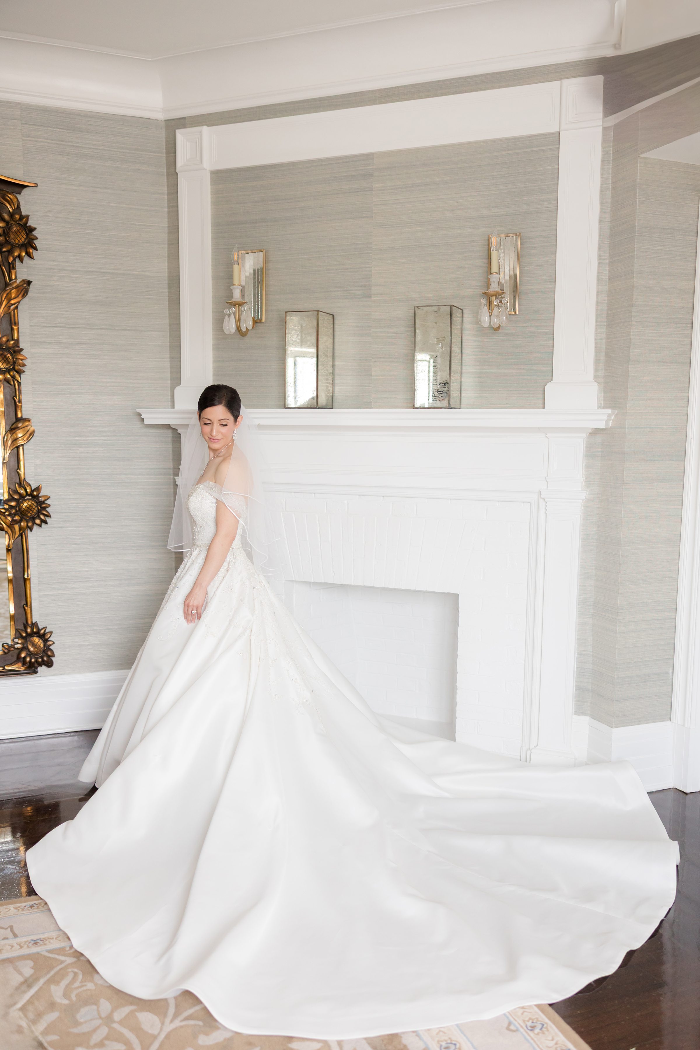 Bride posing by a white fireplace, wearing a short veil and a big wedding dress with a long circular train, looking down