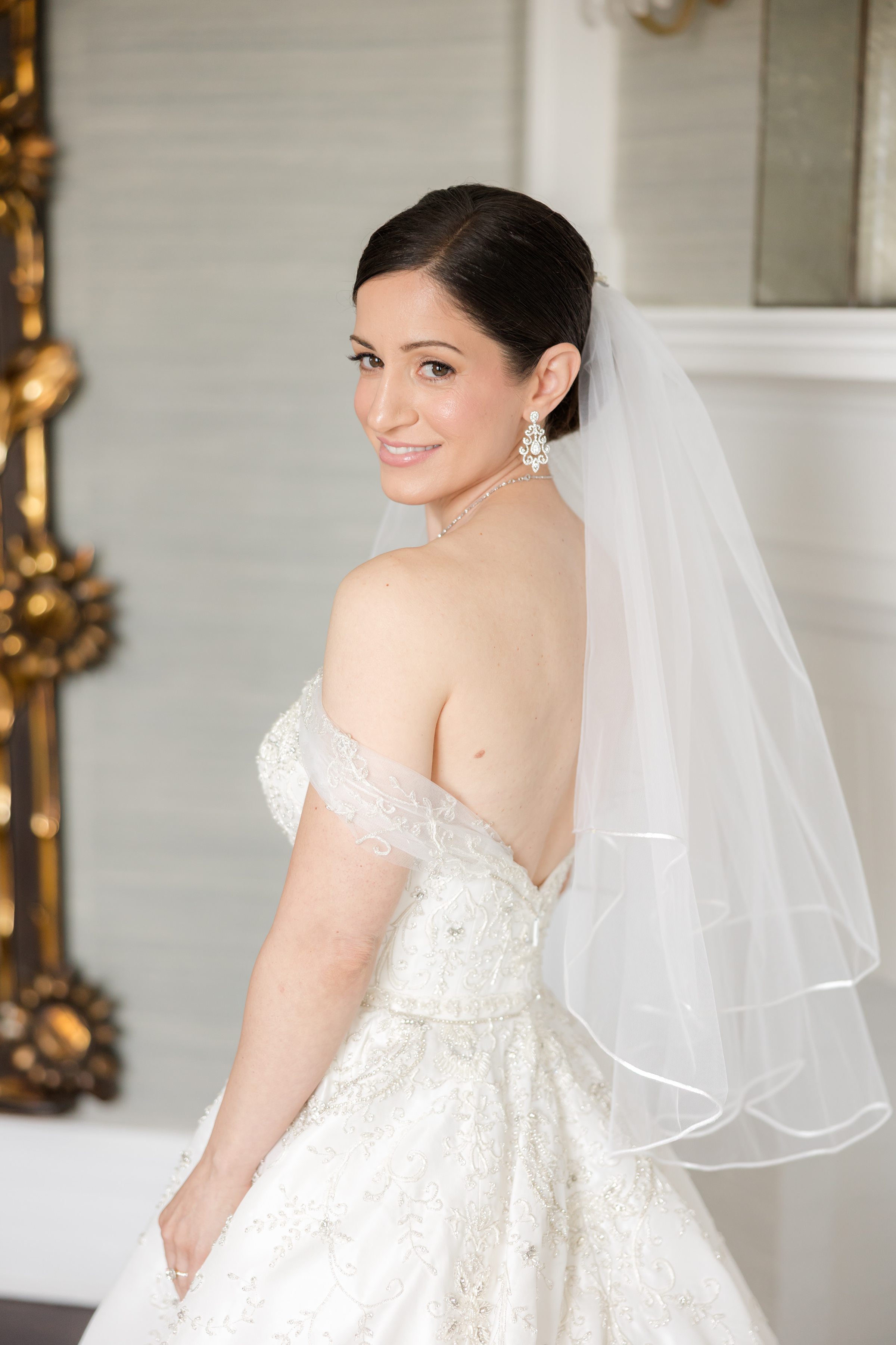 Half-body portrait of bride looking back to the camera, wearing a short veil and an off-shoulder, backless goen. Bride has a birthmark/mole beside her armpit